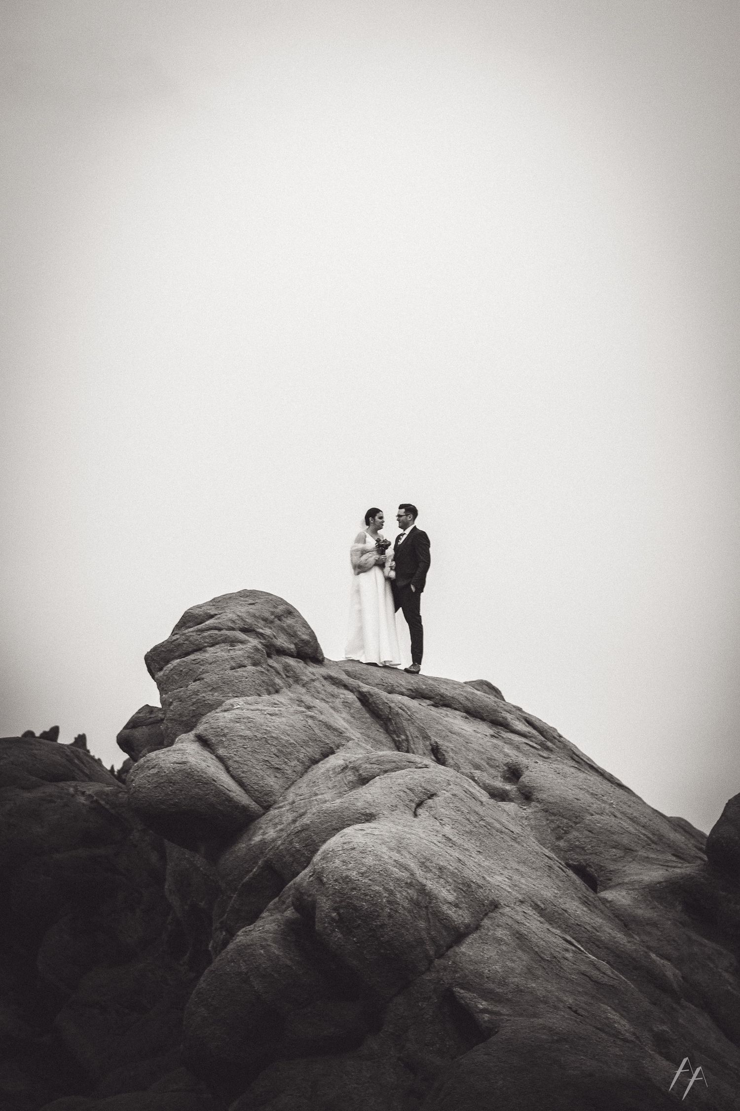 Two silhouettes stand atop a rocky mountain peak against a misty sky in black and white.