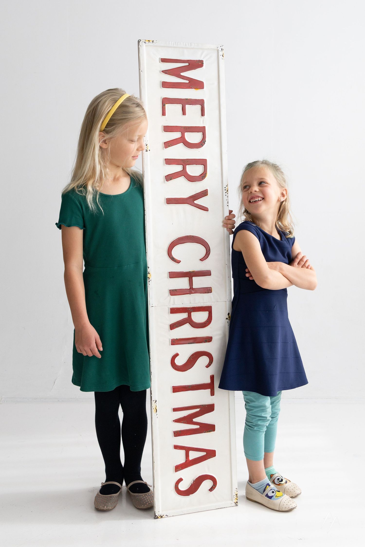 Two small children stand next to a large Merry Christmas sign in a white studio setting.