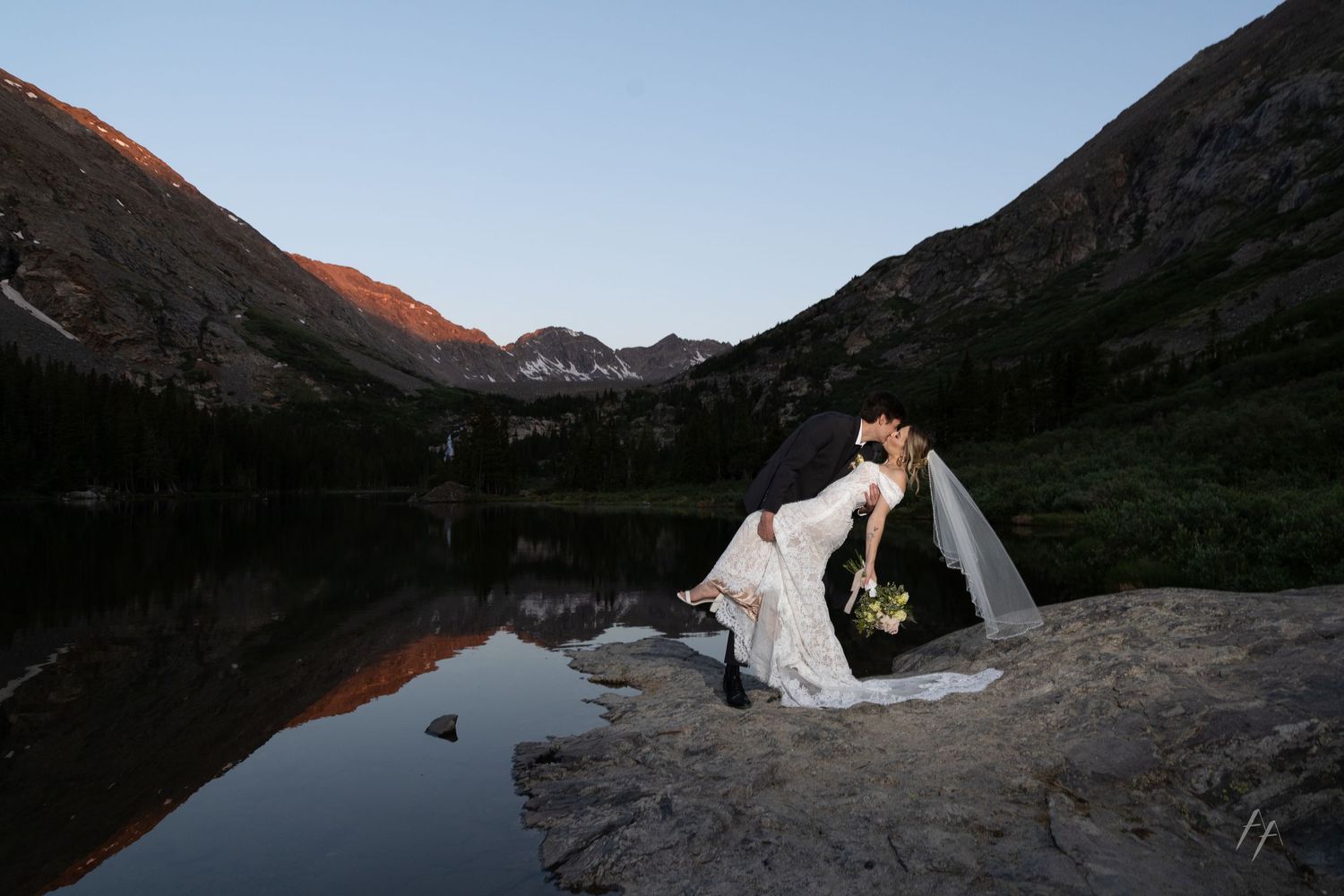 Bride and groom share a romantic kiss on rocks beside a mountain lake at sunset with peaks reflected in calm water.