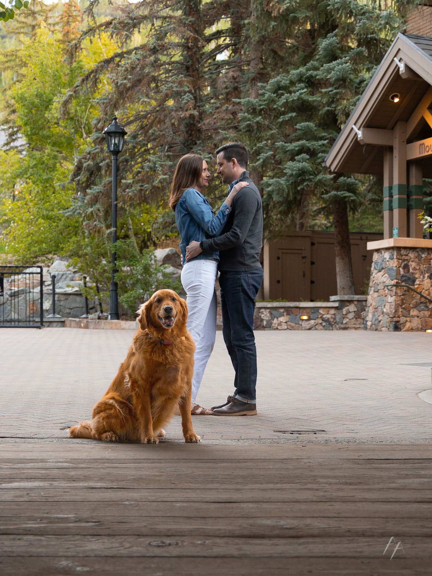 A couple shares a romantic moment in a rustic outdoor setting while their golden retriever sits patiently nearby.