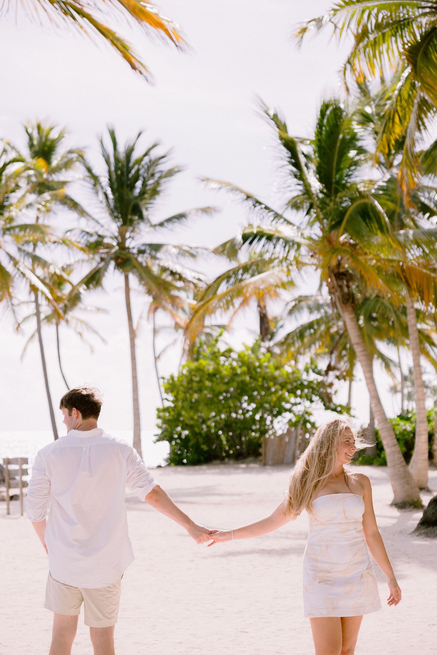 Couple walking hand in hand on a white sandy beach with tall palm trees swaying in the tropical breeze.