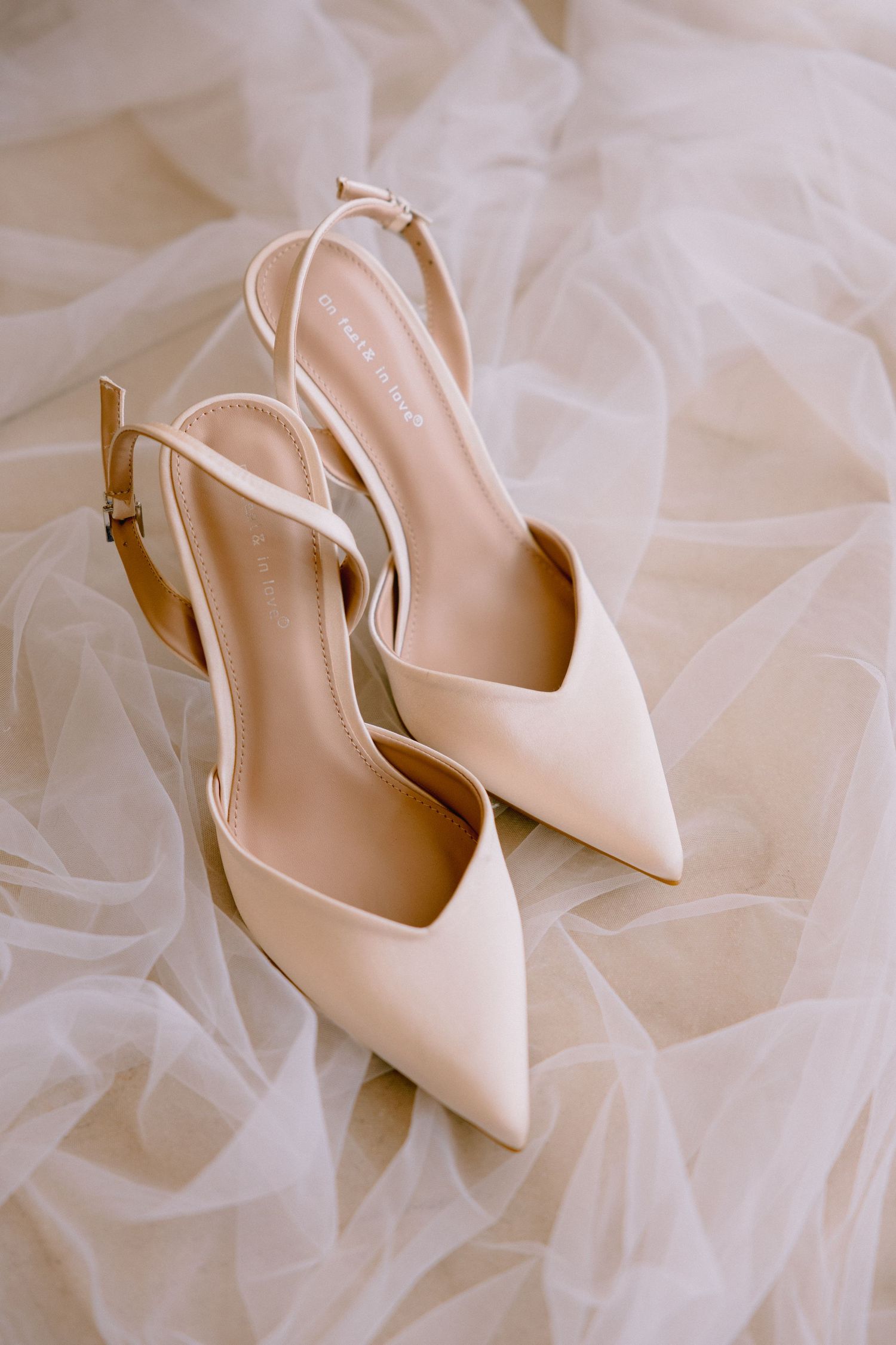 Elegant cream satin slingback heels with pointed toes resting on soft white tulle wedding dress fabric.