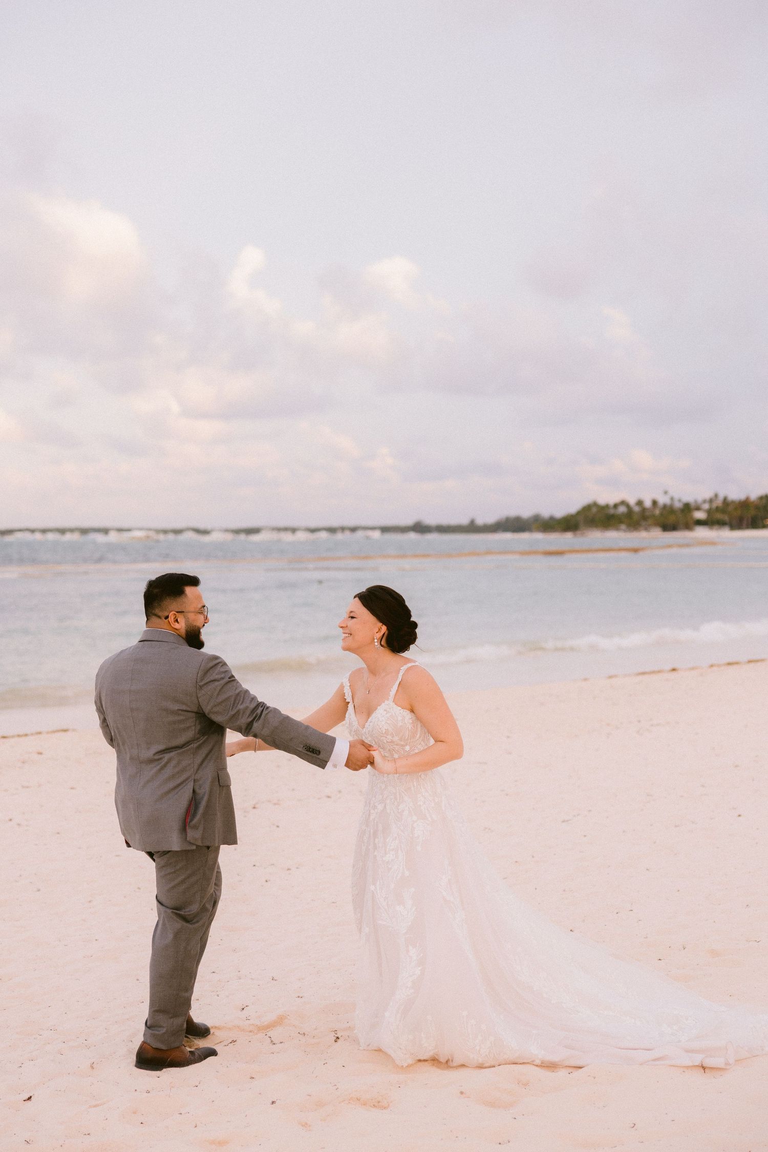 A couple in wedding attire playfully interacts on a serene beach with soft pink sunset lighting.