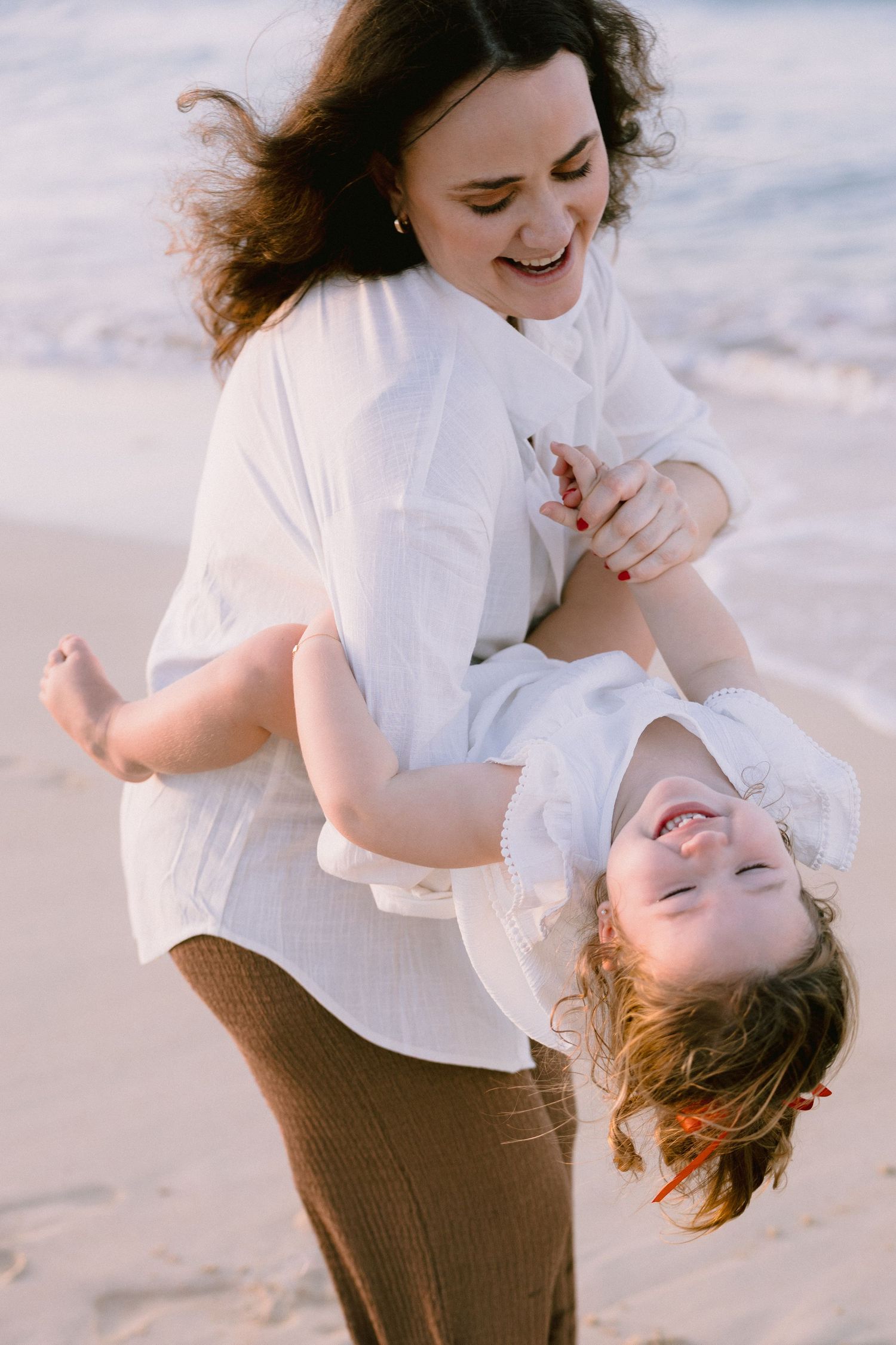 A joyful mother playfully holds her laughing child upside down on a sunny beach.