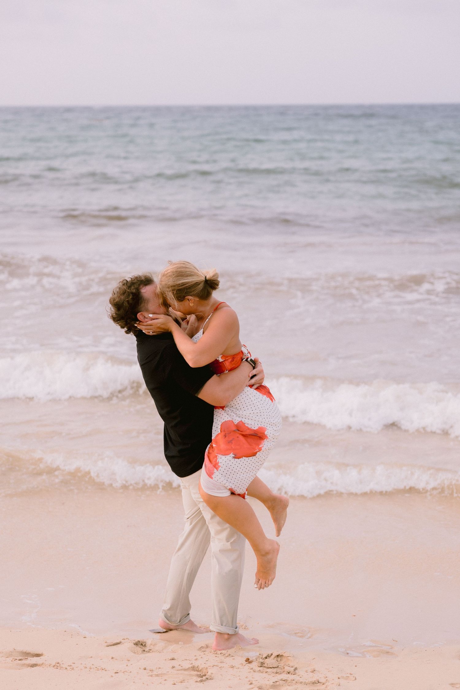 Couple sharing a romantic kiss on a sandy beach, man lifting woman in white and coral dress, ocean waves in background.