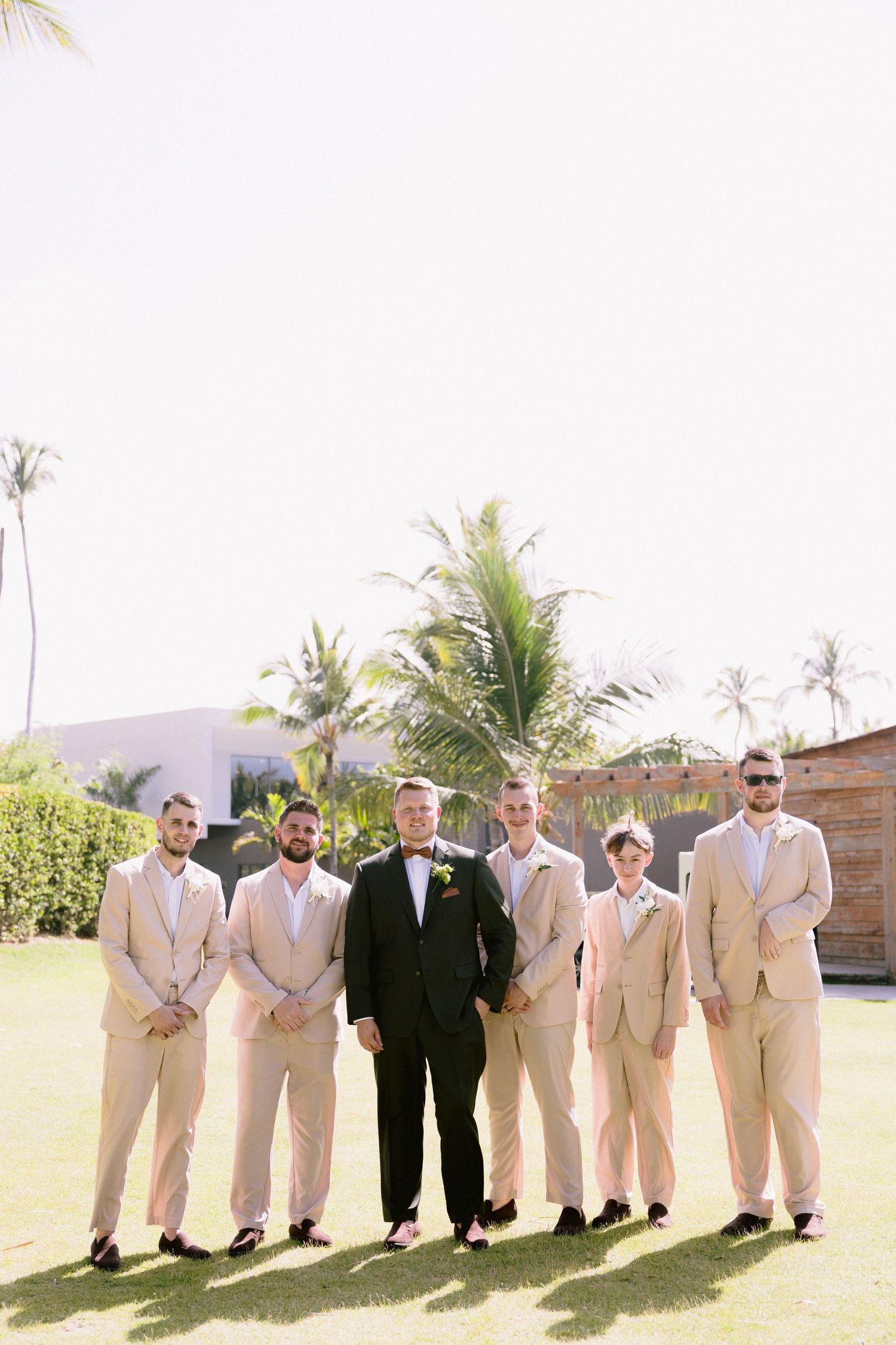 Groom in black tuxedo poses with groomsmen in tan suits on tropical lawn with palm trees.