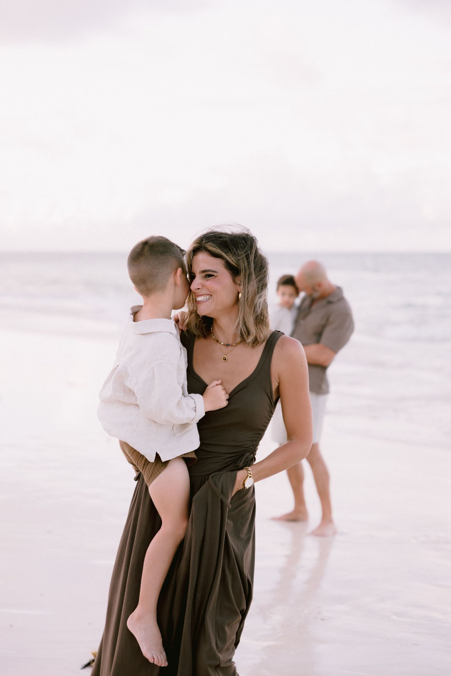 A woman in a brown dress holds a small child while standing on a misty beach with figures in the background.