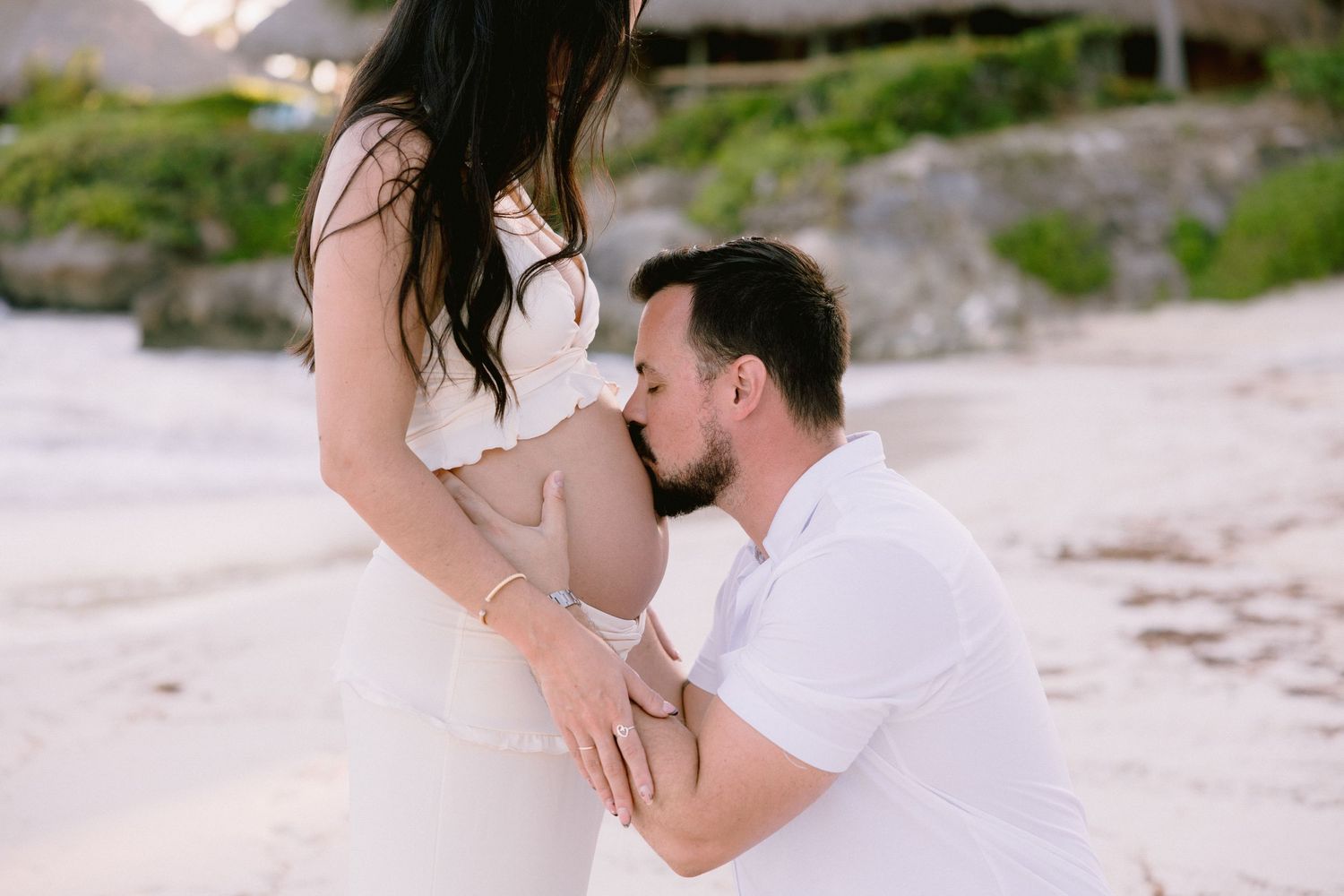 Man in white kneels on beach kissing pregnant woman's belly during a romantic maternity photo shoot.