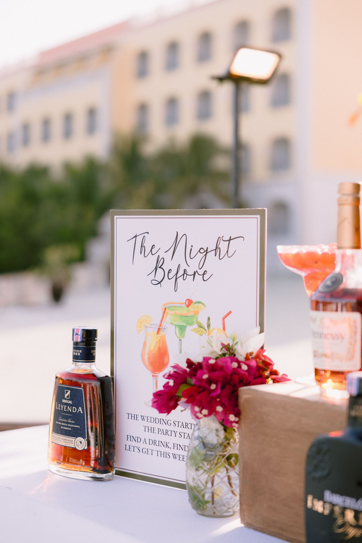 Welcome sign reading The Night Before beside pink flowers and liquor bottles on a table at an outdoor wedding welcome party.