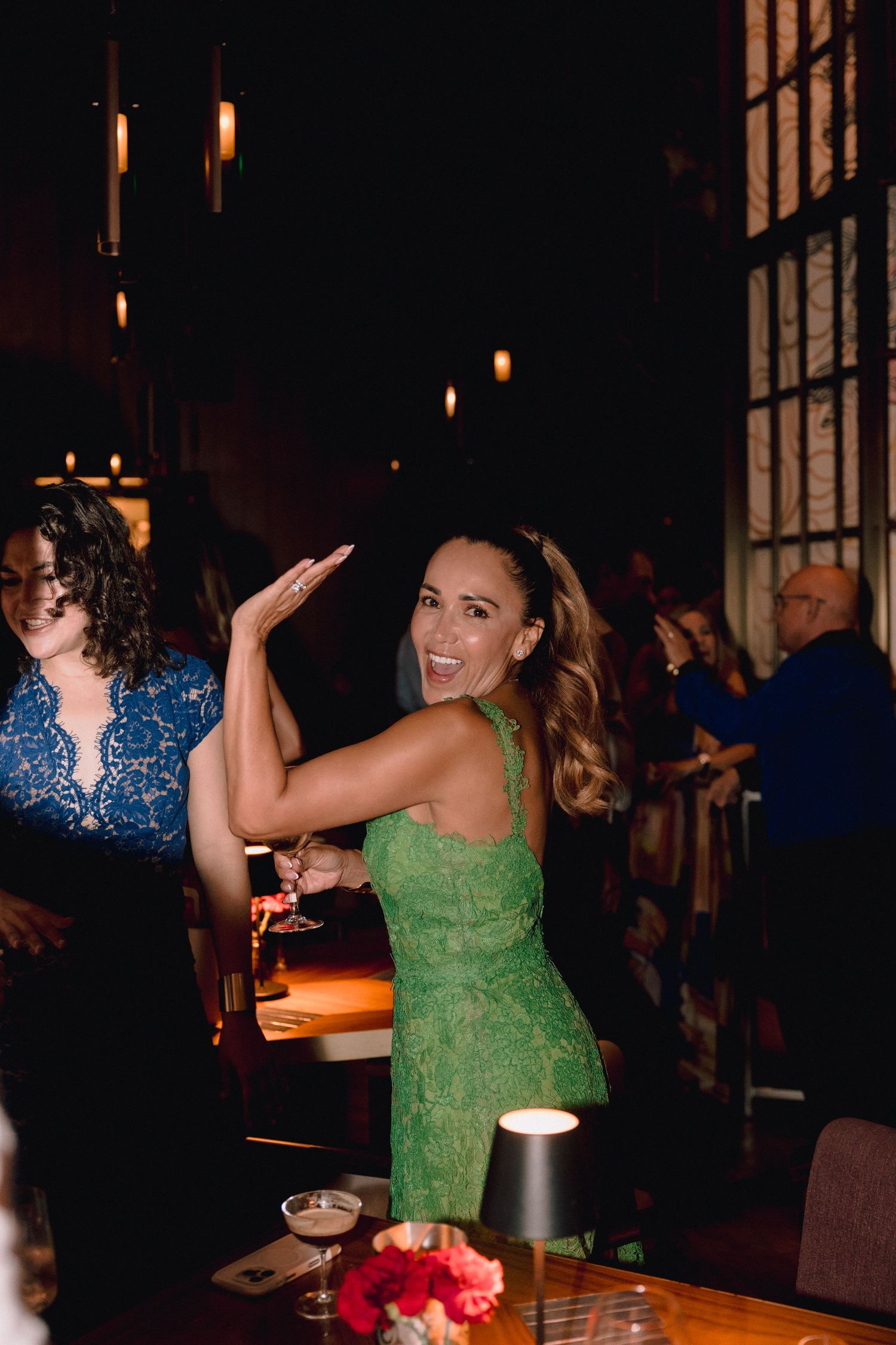 Smiling woman in a green lace dress dances with a cocktail in hand at a dimly lit restaurant, with a rose centerpiece and guests nearby.