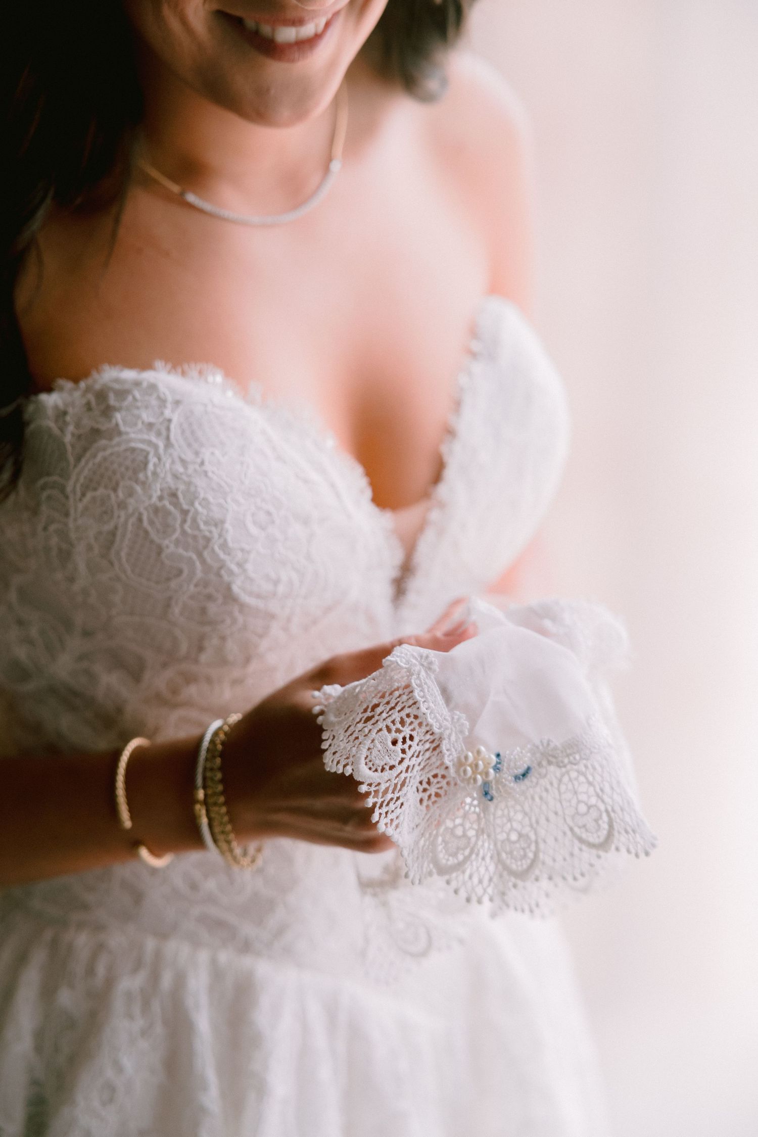 Smiling bride in strapless lace wedding gown holding delicate white lace garter with blue details and pearl accents.