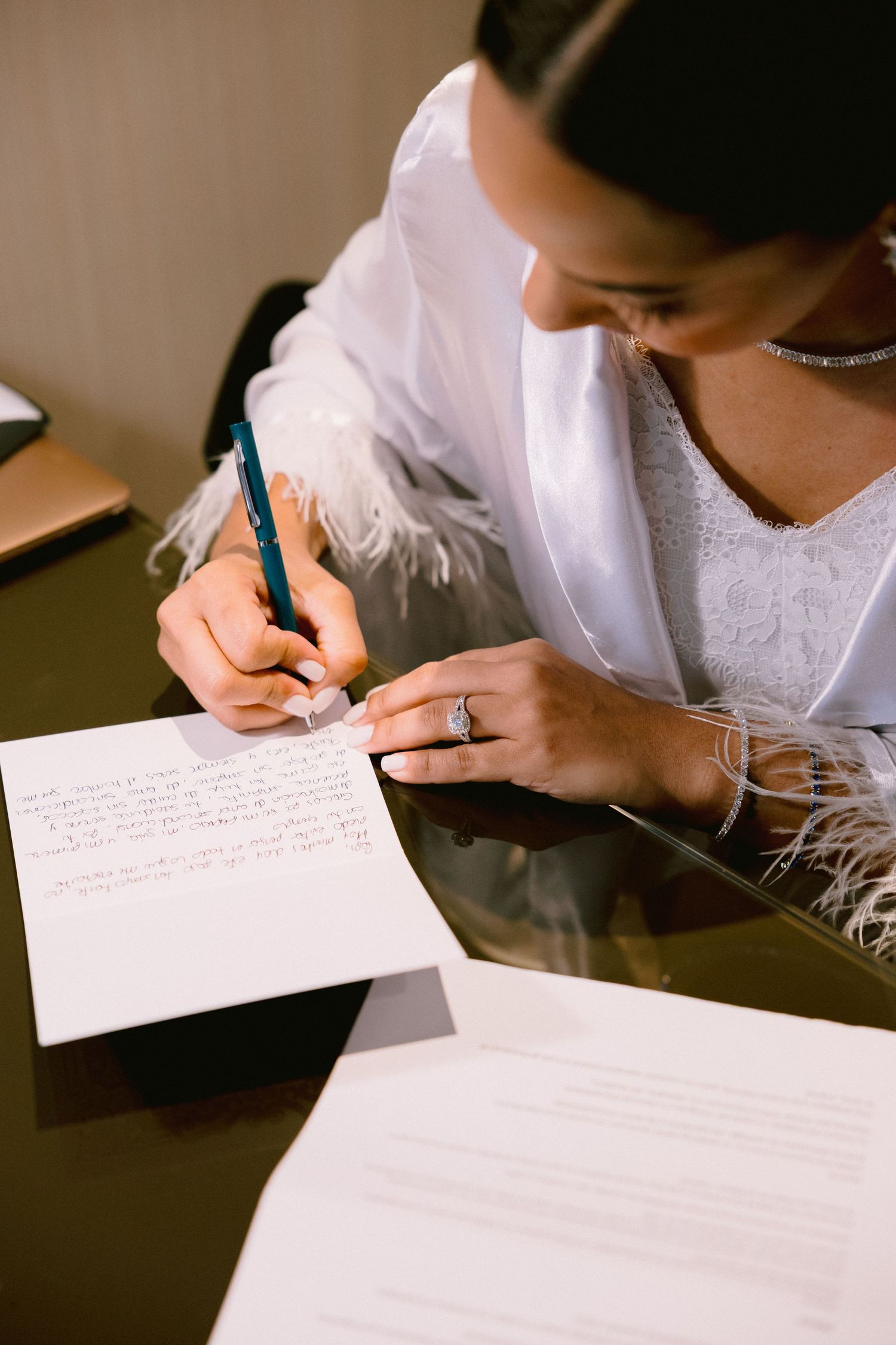 Bride in white feathered robe writing heartfelt vows at desk, wearing diamond ring and necklace.
