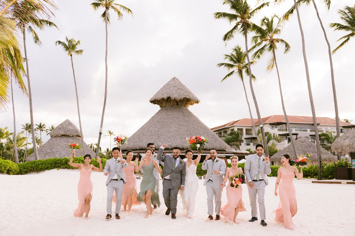 Wedding party walks along a tropical beach with palm trees and thatched roof huts in the background.