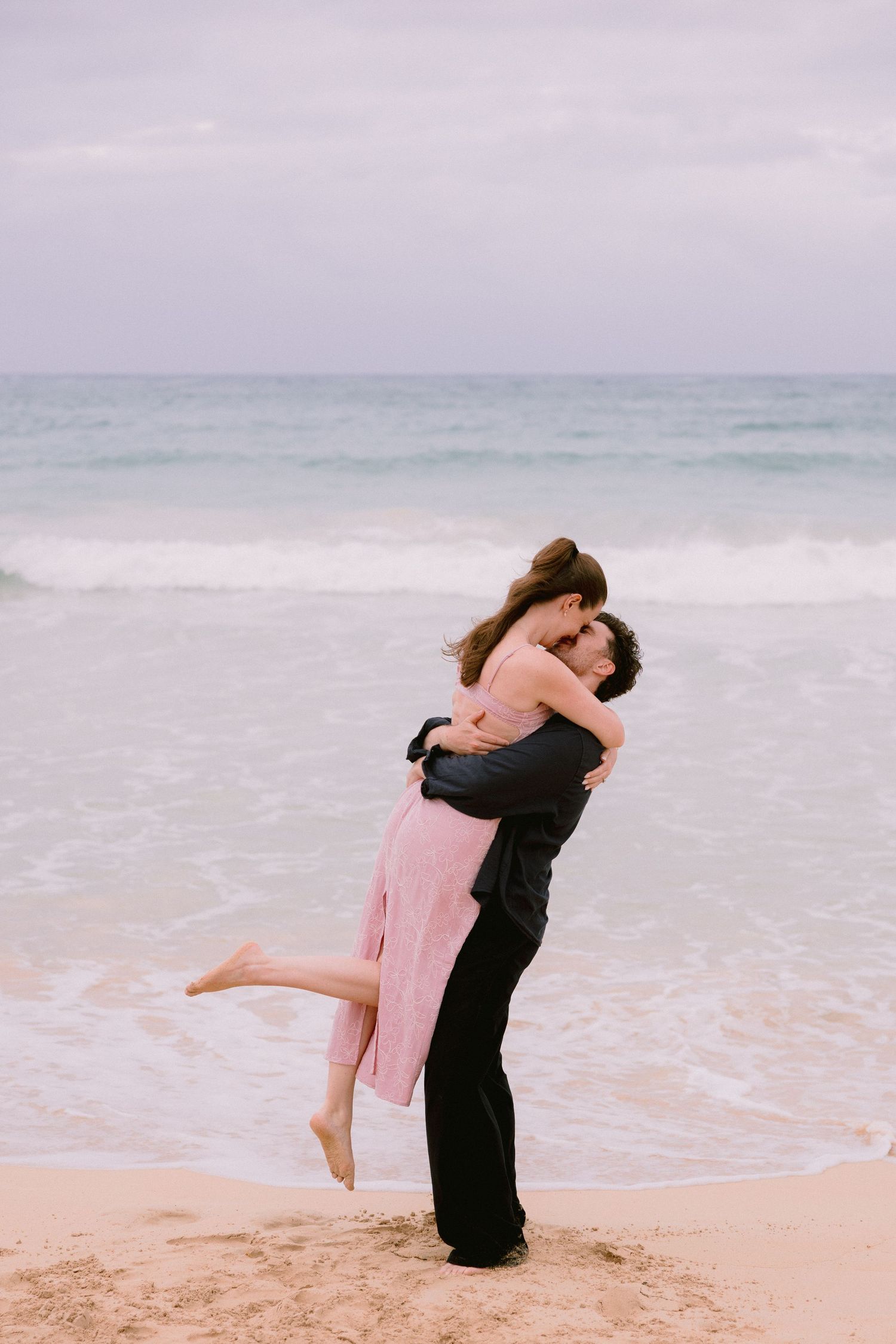 Couple embracing and kissing on a sandy beach with ocean waves, man lifting woman in pink dress.