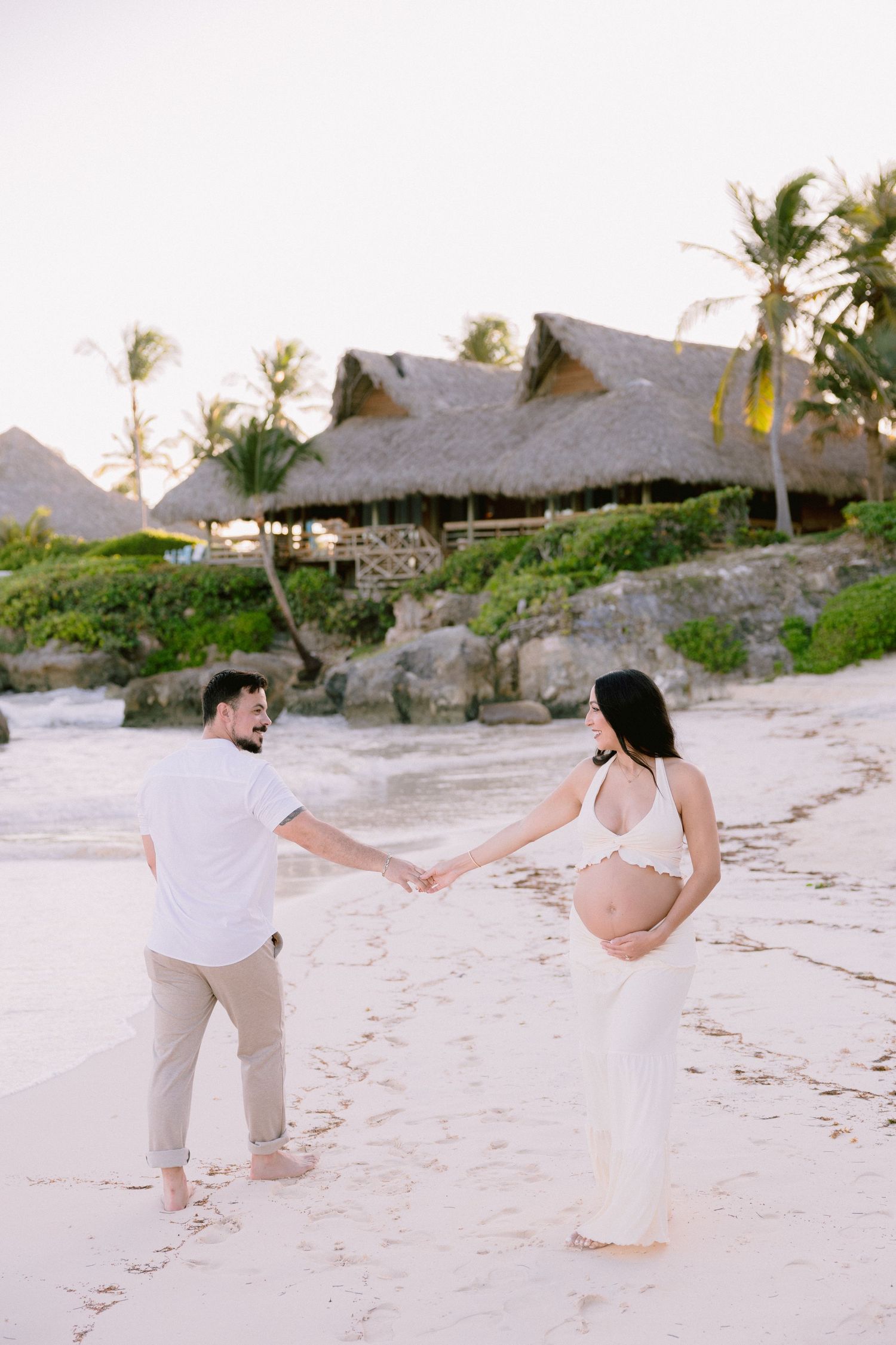 Pregnant couple holding hands on tropical beach with palm trees and thatched huts, maternity photoshoot at sunset.