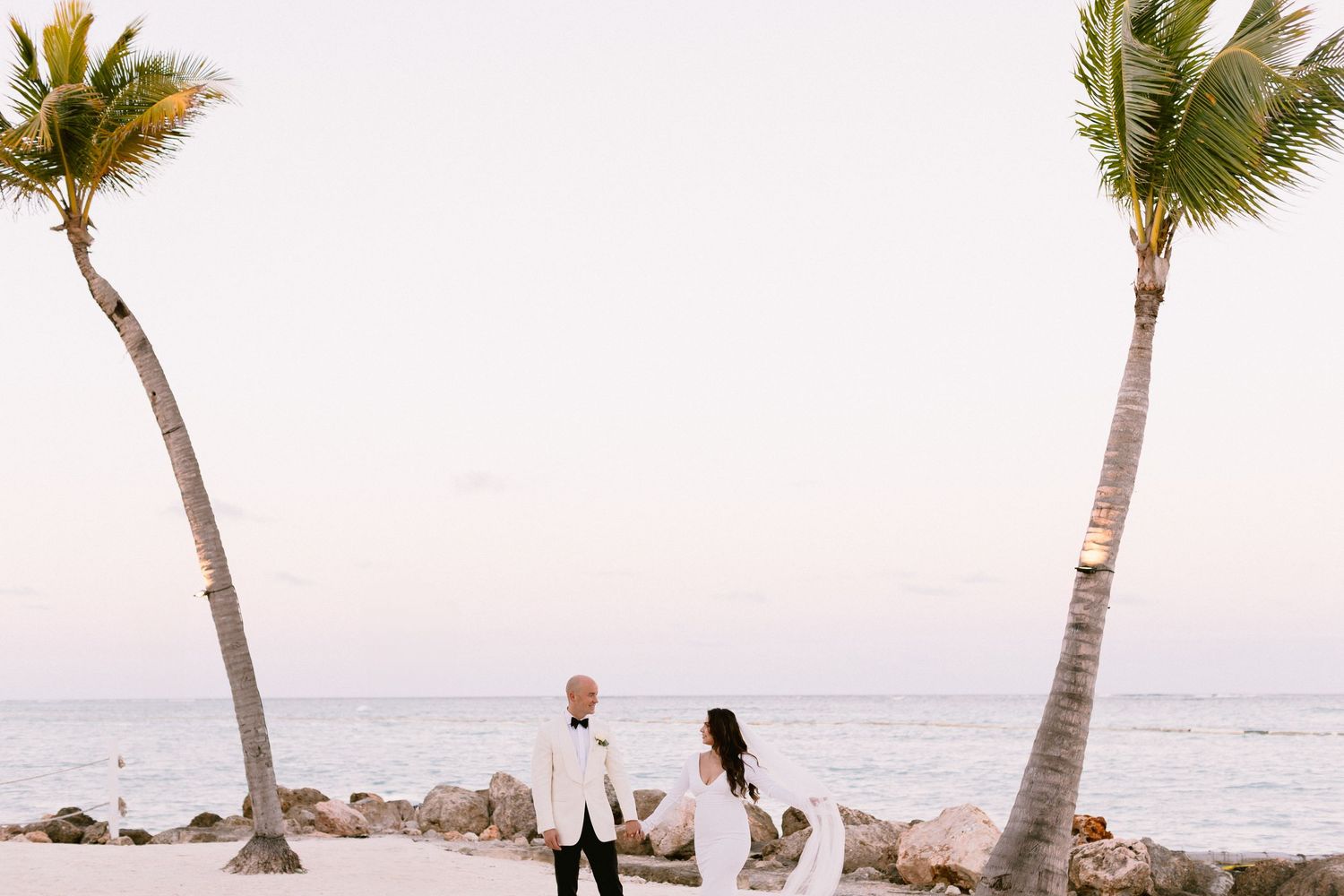 Couple holding hands on a tropical beach wedding with palm trees and ocean backdrop at sunset.