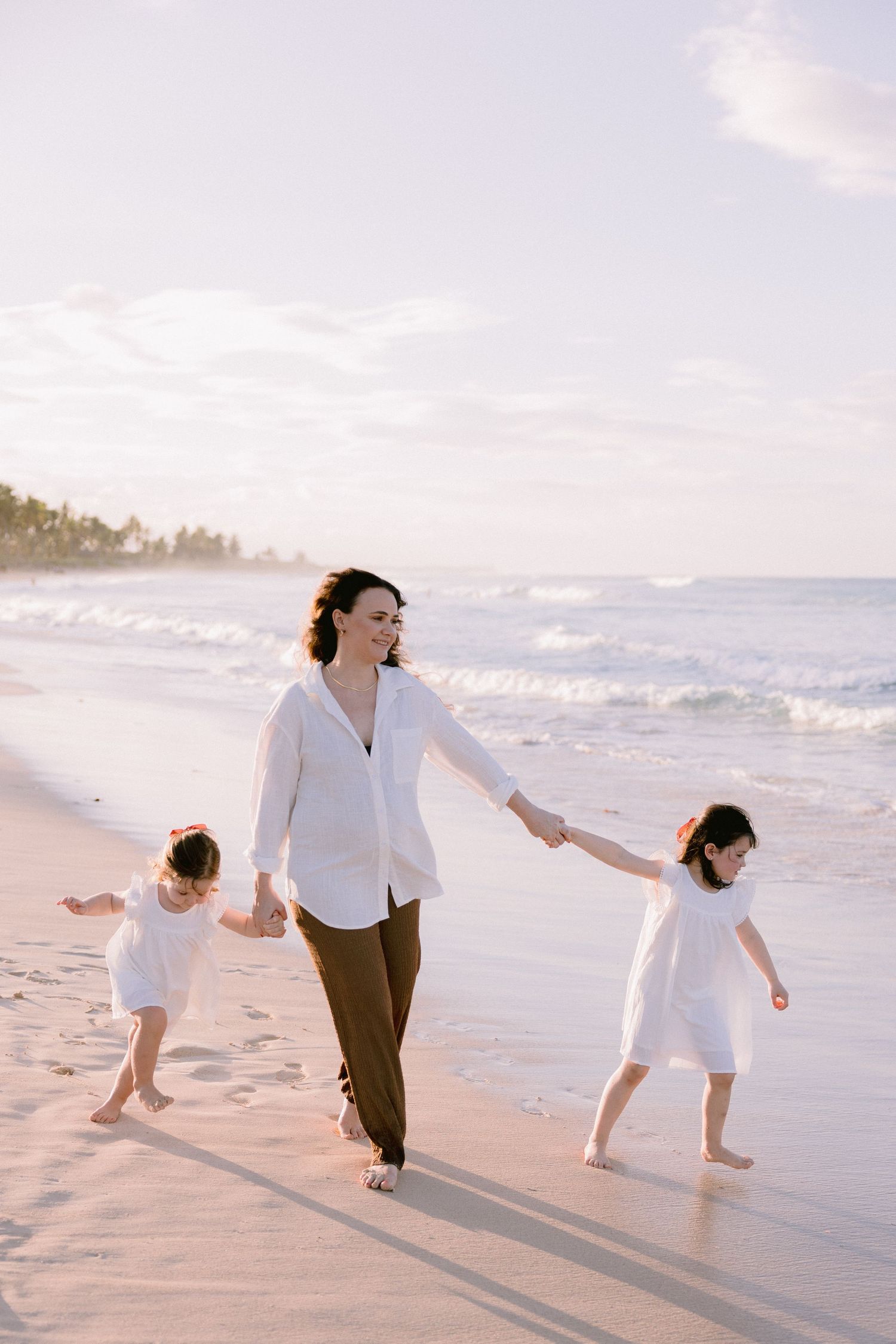 Two people in white and brown outfits playfully dance together on a sunlit beach at sunset.
