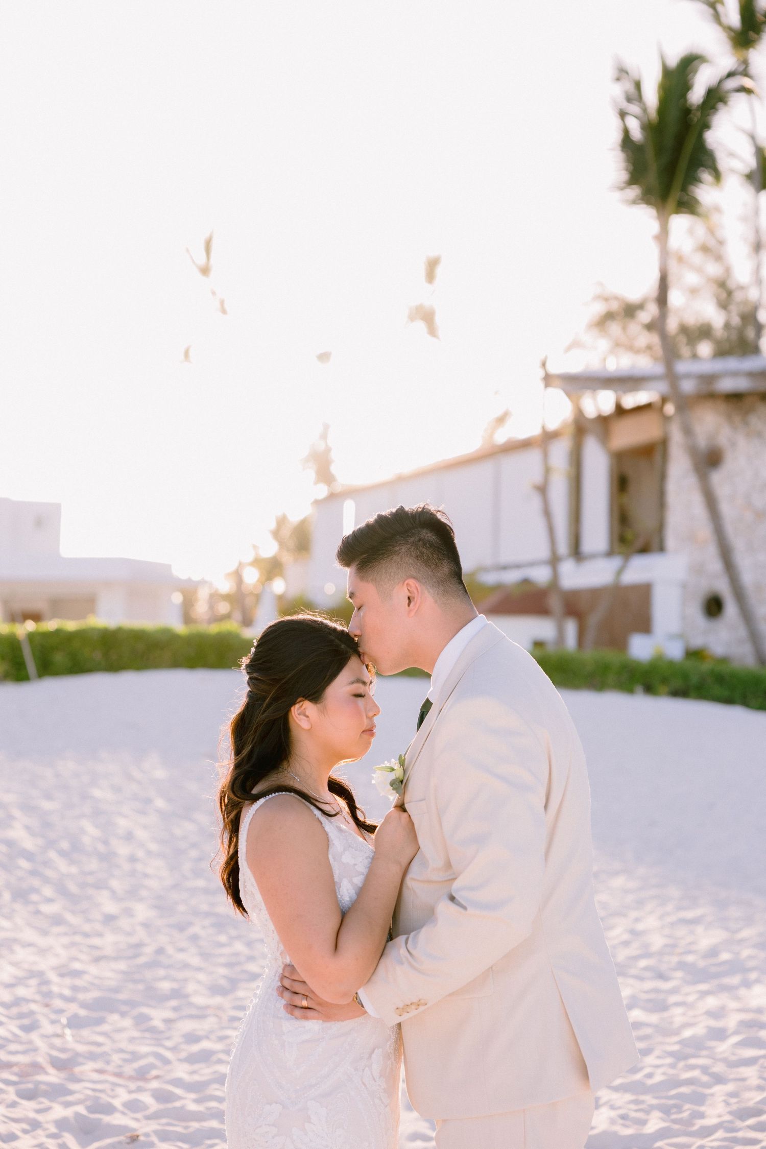 Bride and groom share a romantic moment on a sandy beach at sunset, with the groom kissing her forehead.