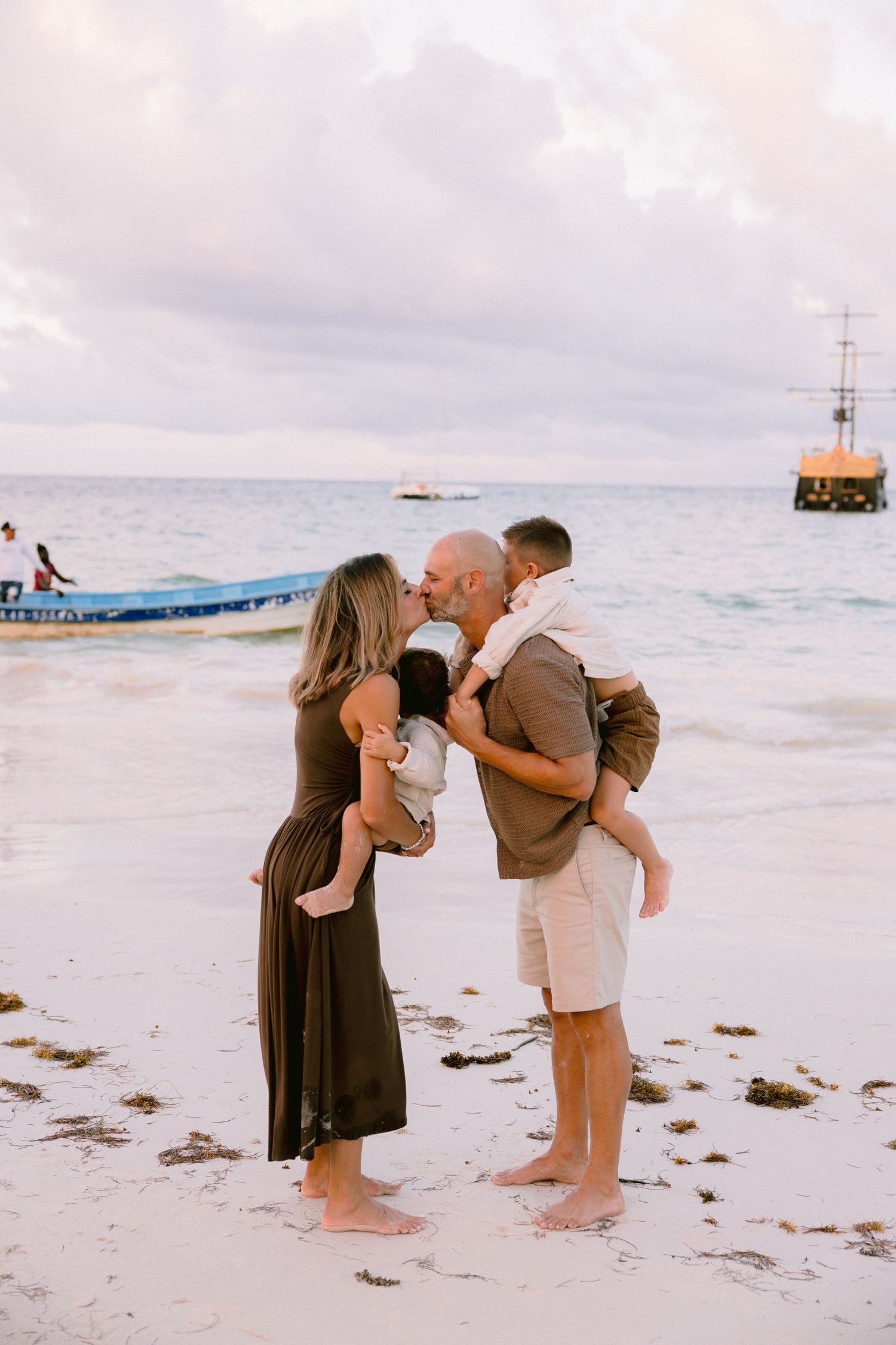 Couple kissing on a tropical beach at sunset, each holding a young child, with a calm ocean and boats in the background.