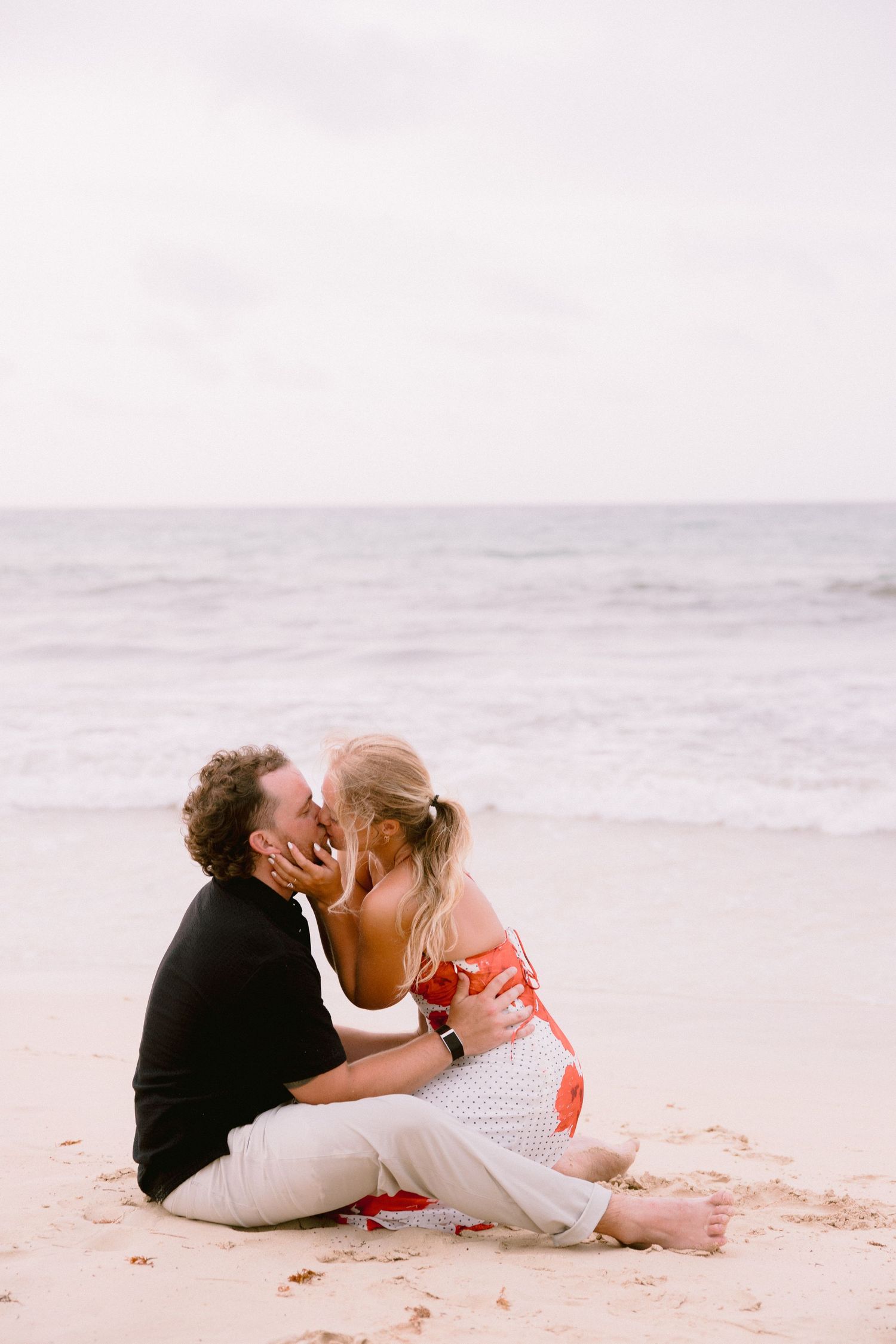 Couple kissing romantically on a sandy beach, woman in red dress and man in black shirt, with ocean waves in background.