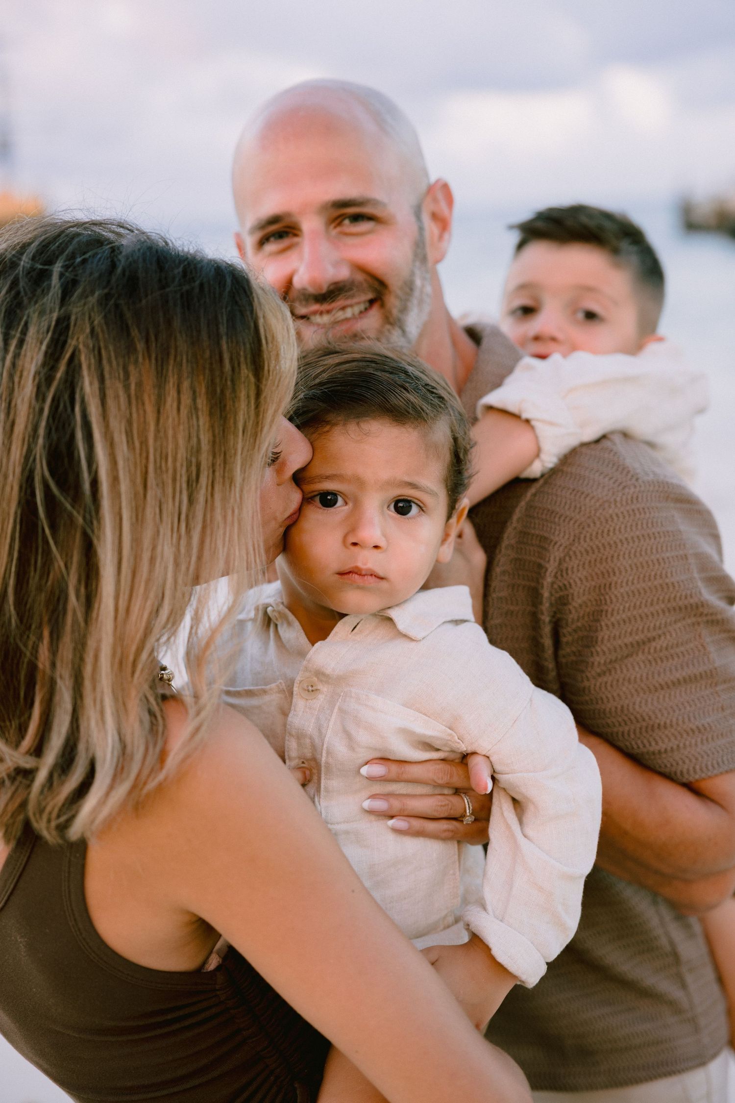 A smiling bald man holds two young children while a blonde woman kisses one child on a beach at sunset.