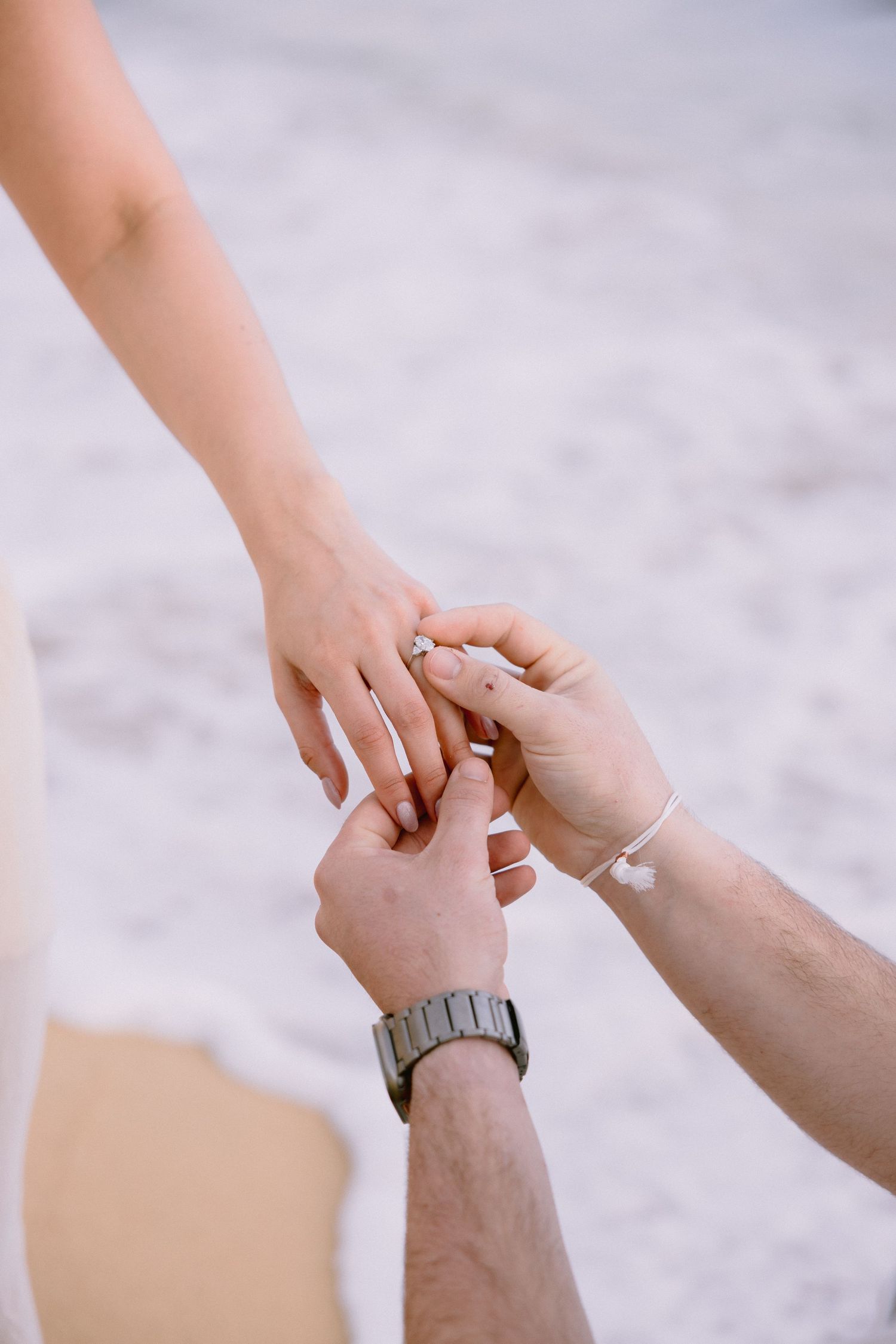 Man placing an engagement ring on woman's finger on a sandy beach, romantic proposal moment.