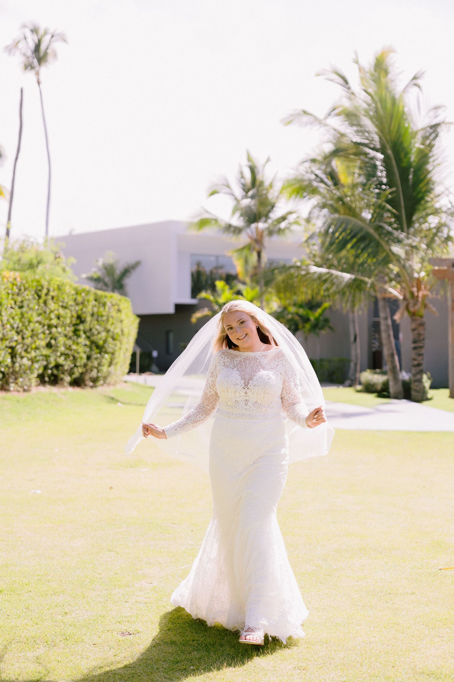 Bride in lace mermaid gown and veil walks on green lawn with tropical palm trees in sunny destination wedding.