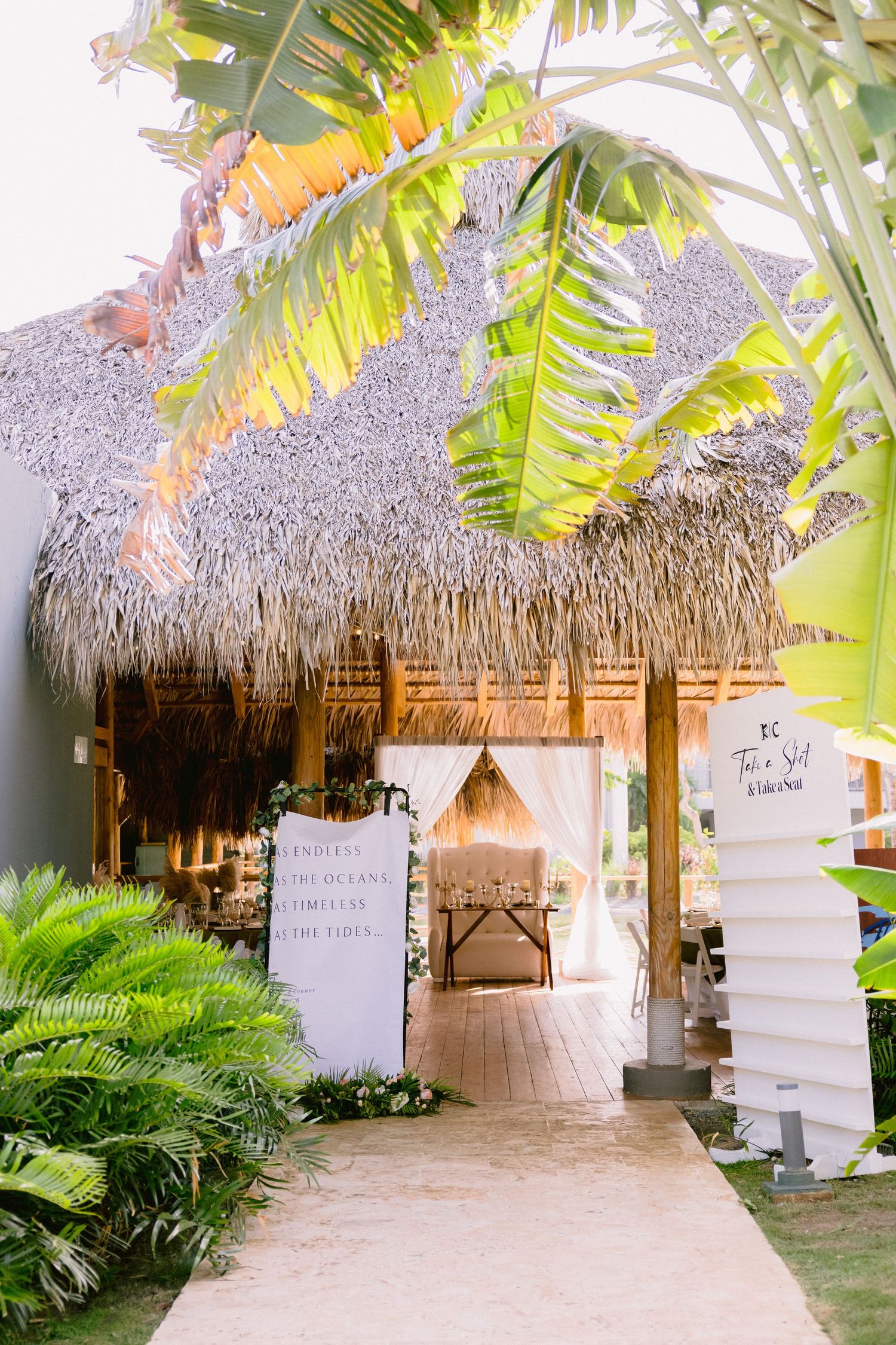 Tropical wedding venue entrance with thatched roof palapa, lush palm trees, and elegant white decor along a sandy pathway.