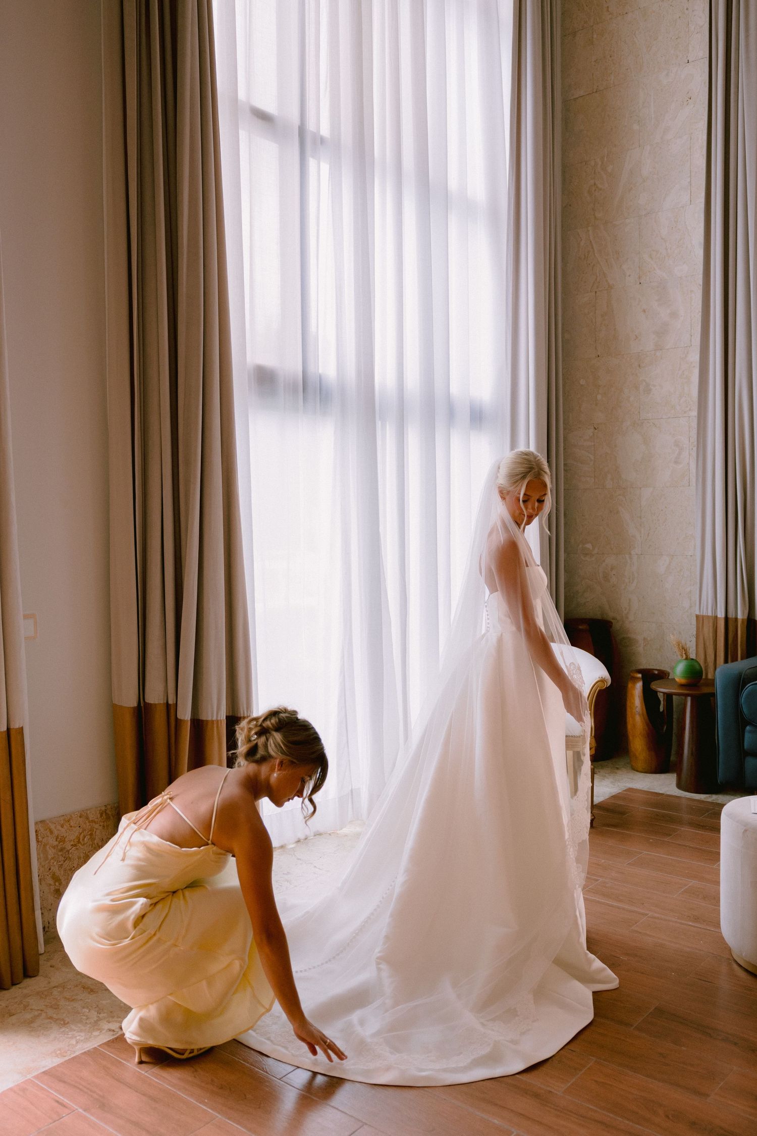 Bridesmaid in yellow dress adjusting the train of a brides white wedding gown near a bright window.
