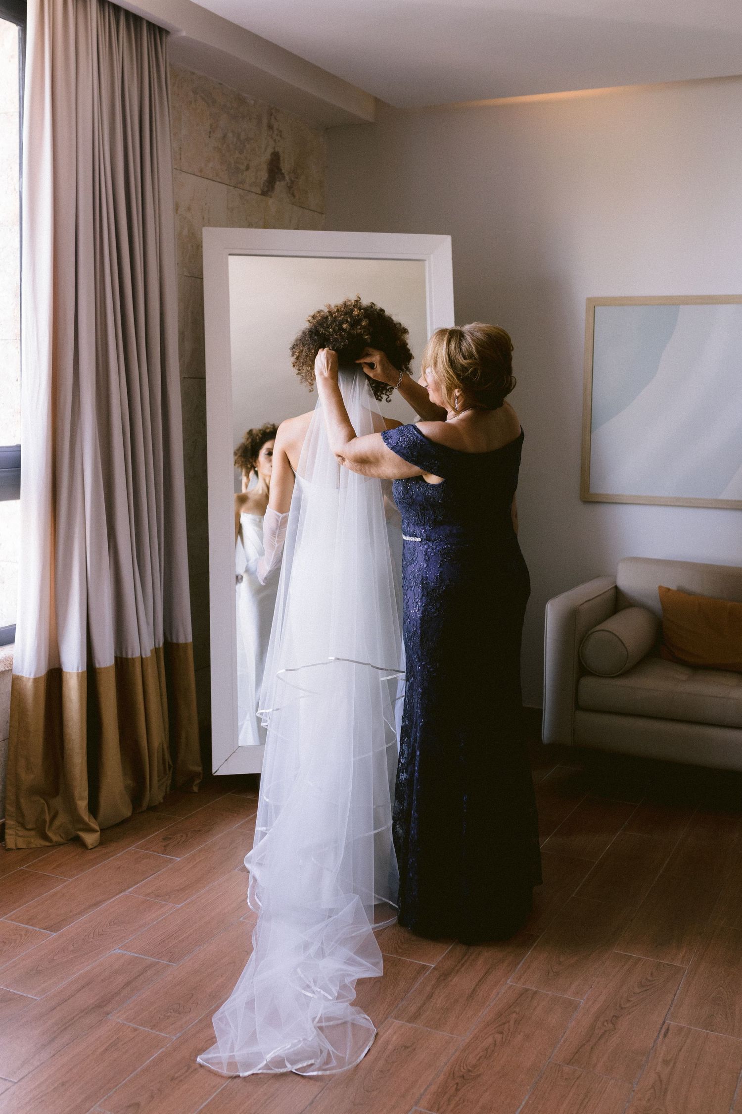 A woman in a blue gown helps a bride in a white wedding dress and veil prepare in a hotel room.