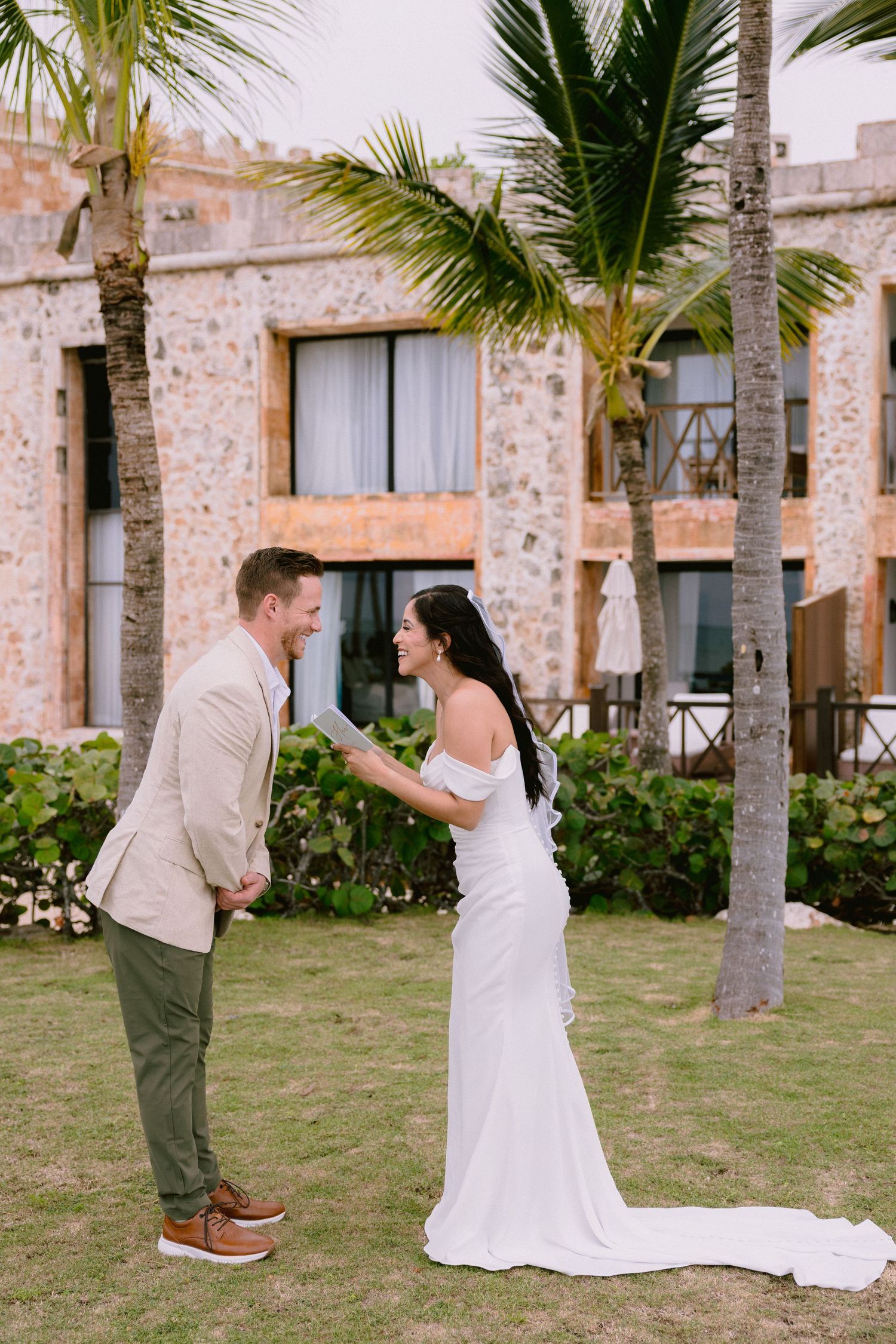 Bride reads vows to groom in white off-shoulder gown at tropical resort wedding ceremony with palm trees.