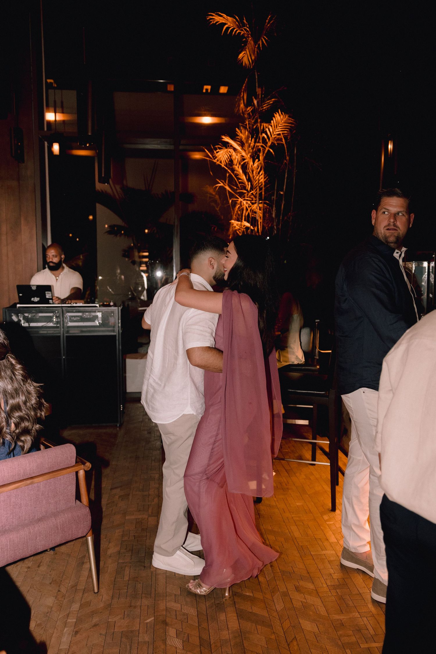 A couple embraces and dances closely on a wood floor in a dimly lit lounge with warm lighting, a DJ booth, and guests nearby.