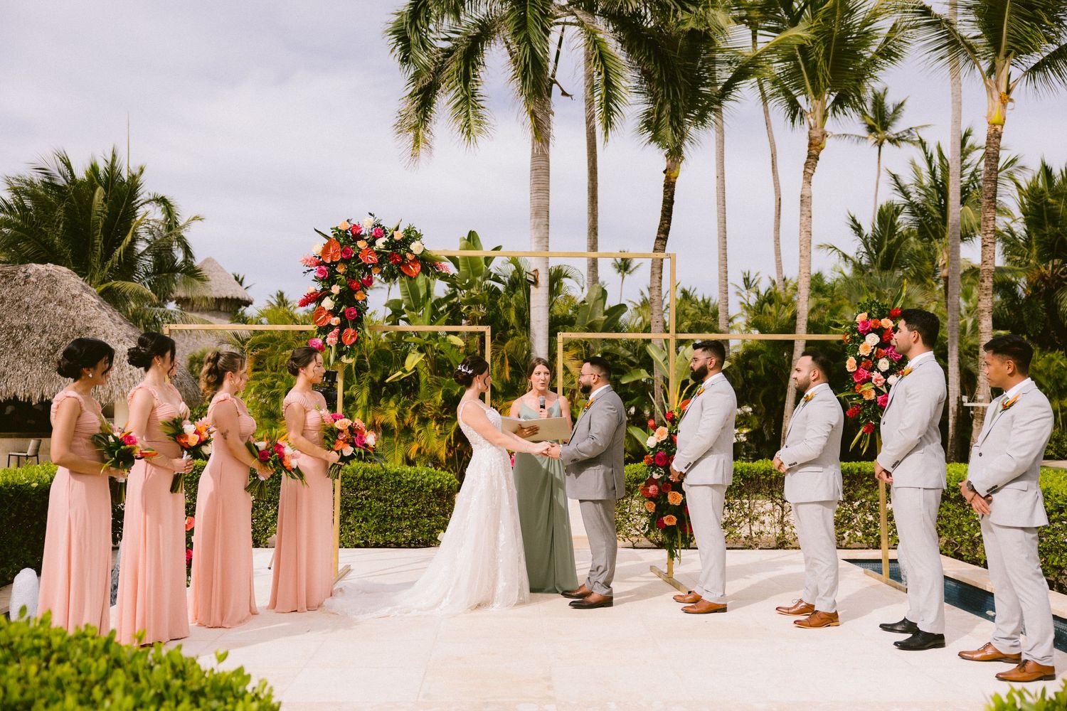 Wedding party in blush pink and white attire stands in line against a backdrop of tropical palm trees at sunset.