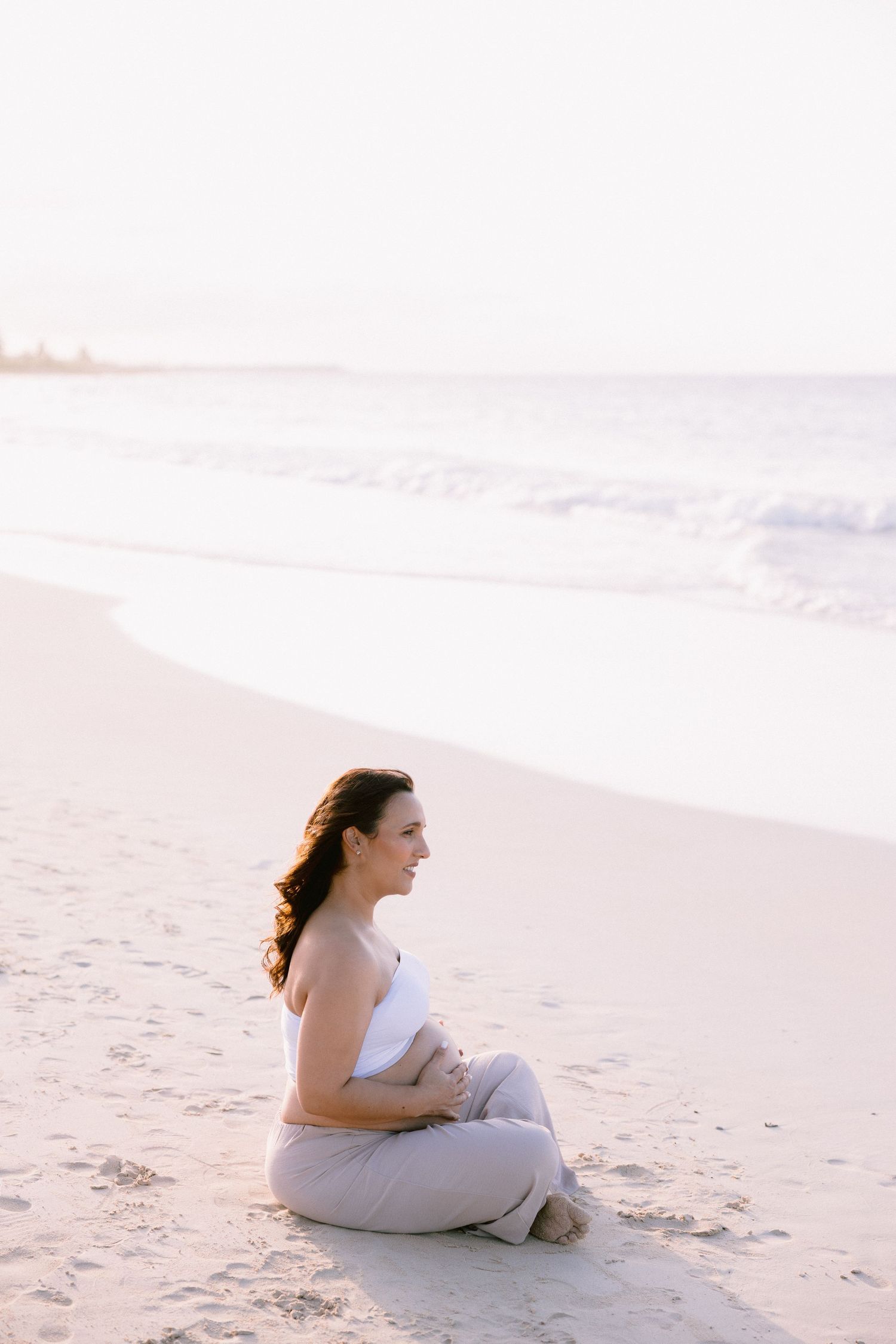 A woman sits cross-legged on a sandy beach, gazing at ocean waves during a serene golden hour sunset.
