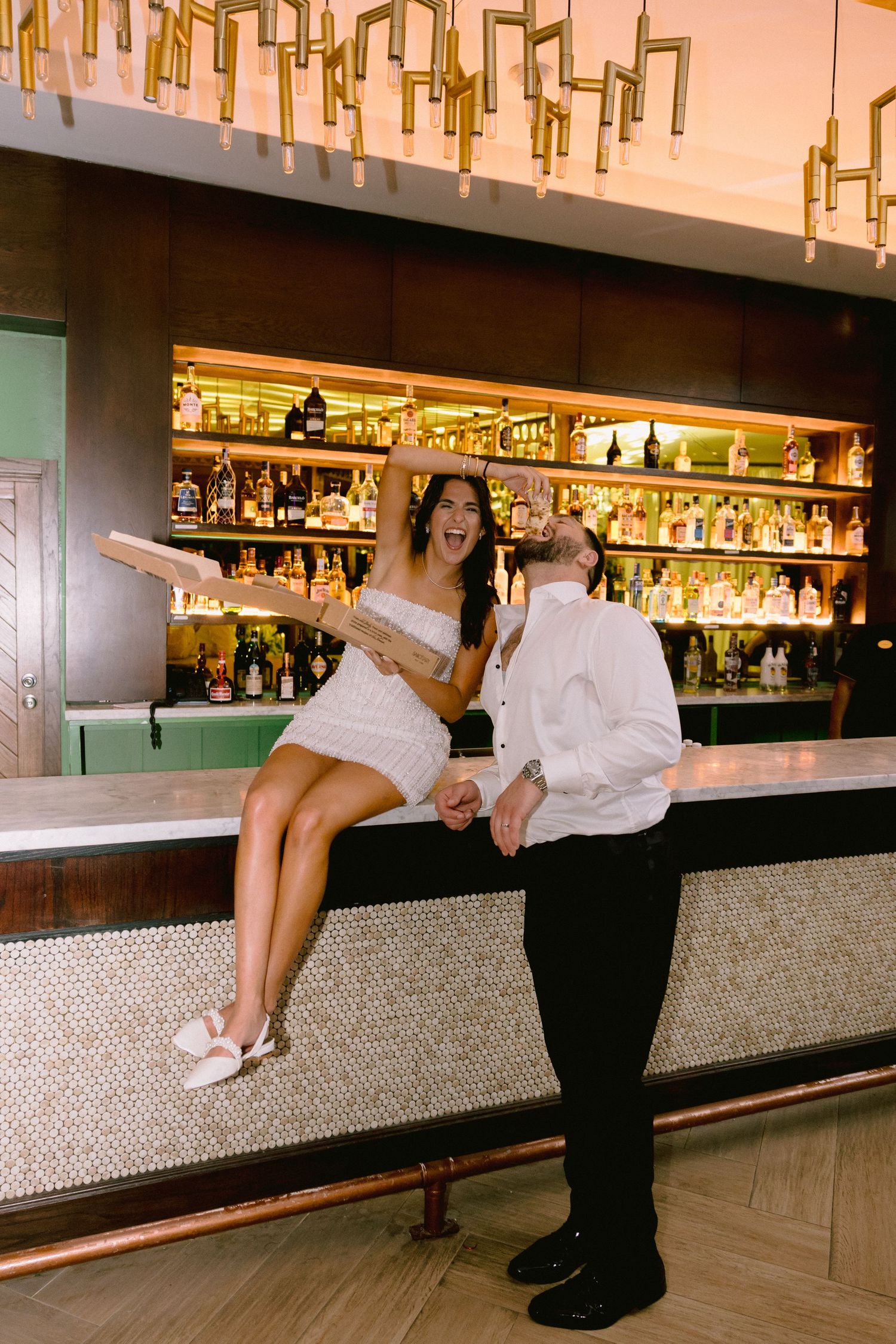 A laughing bride in a white mini dress sits on a bar counter holding hands with a groom in a white shirt.