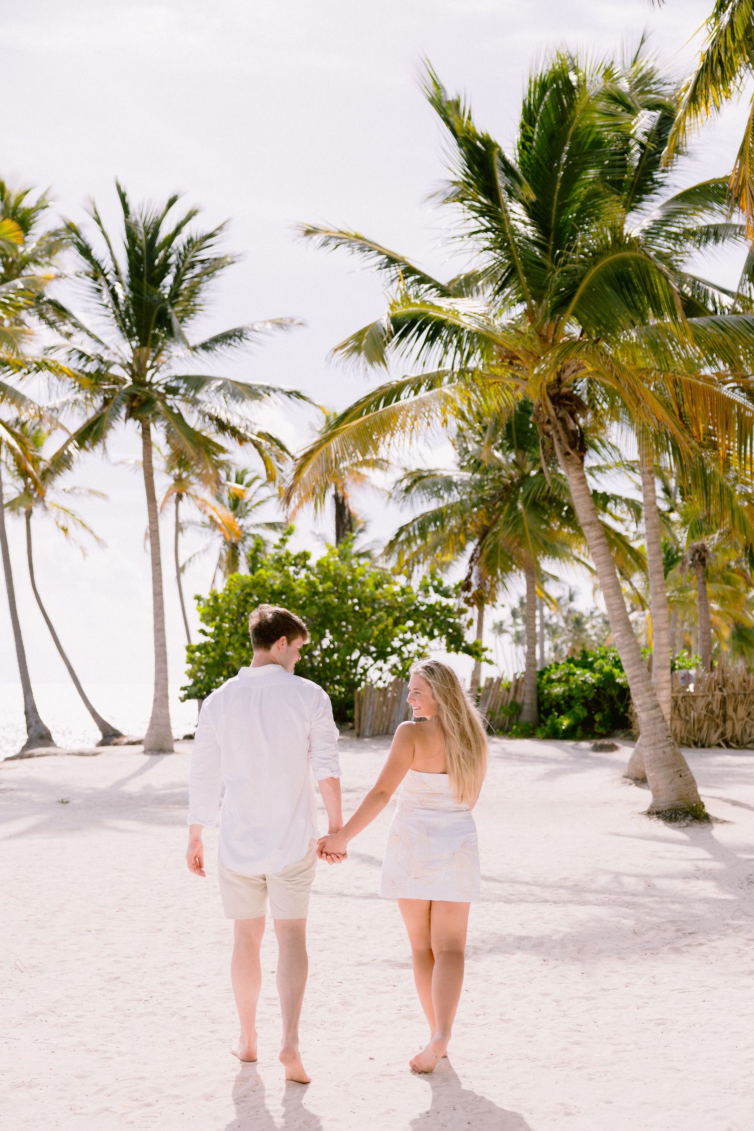 Couple walking hand in hand on a white sandy beach with tall palm trees lining the tropical shoreline.