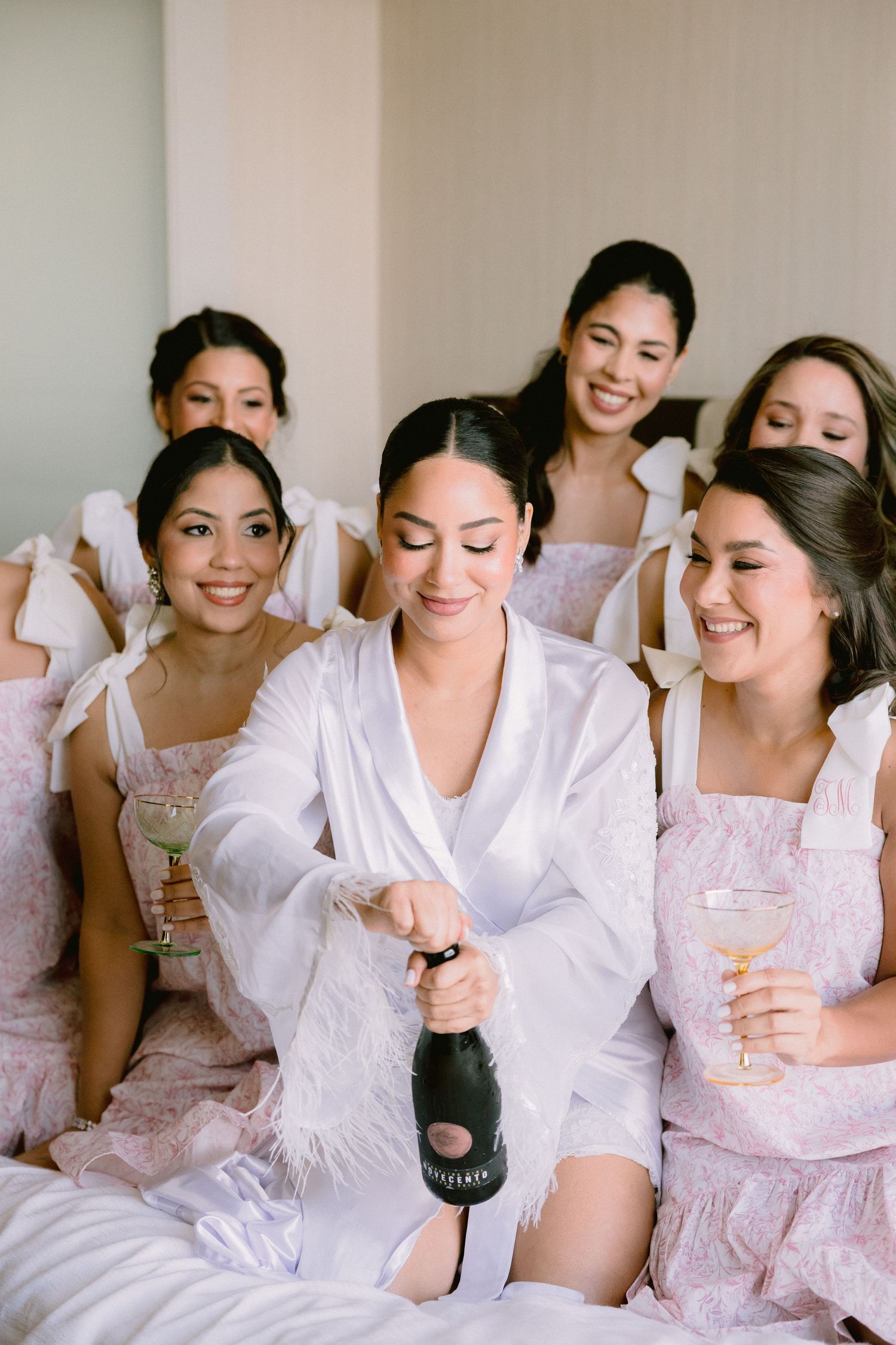 Bride in white feather robe opens champagne bottle surrounded by bridesmaids in pink floral robes during wedding morning prep.
