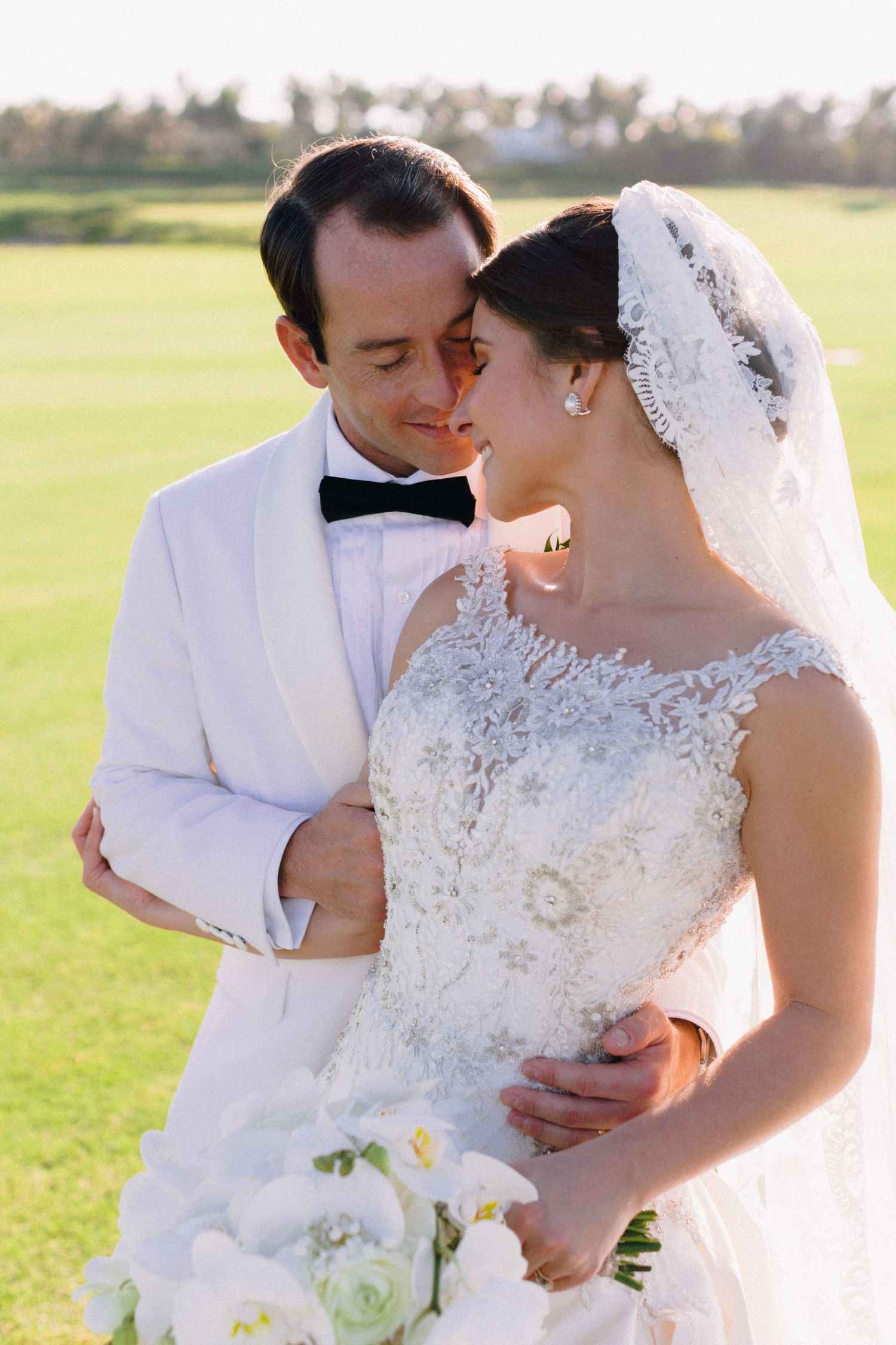 A romantic wedding couple shares intimate moments in a sunlit field while holding a white and cream bouquet.