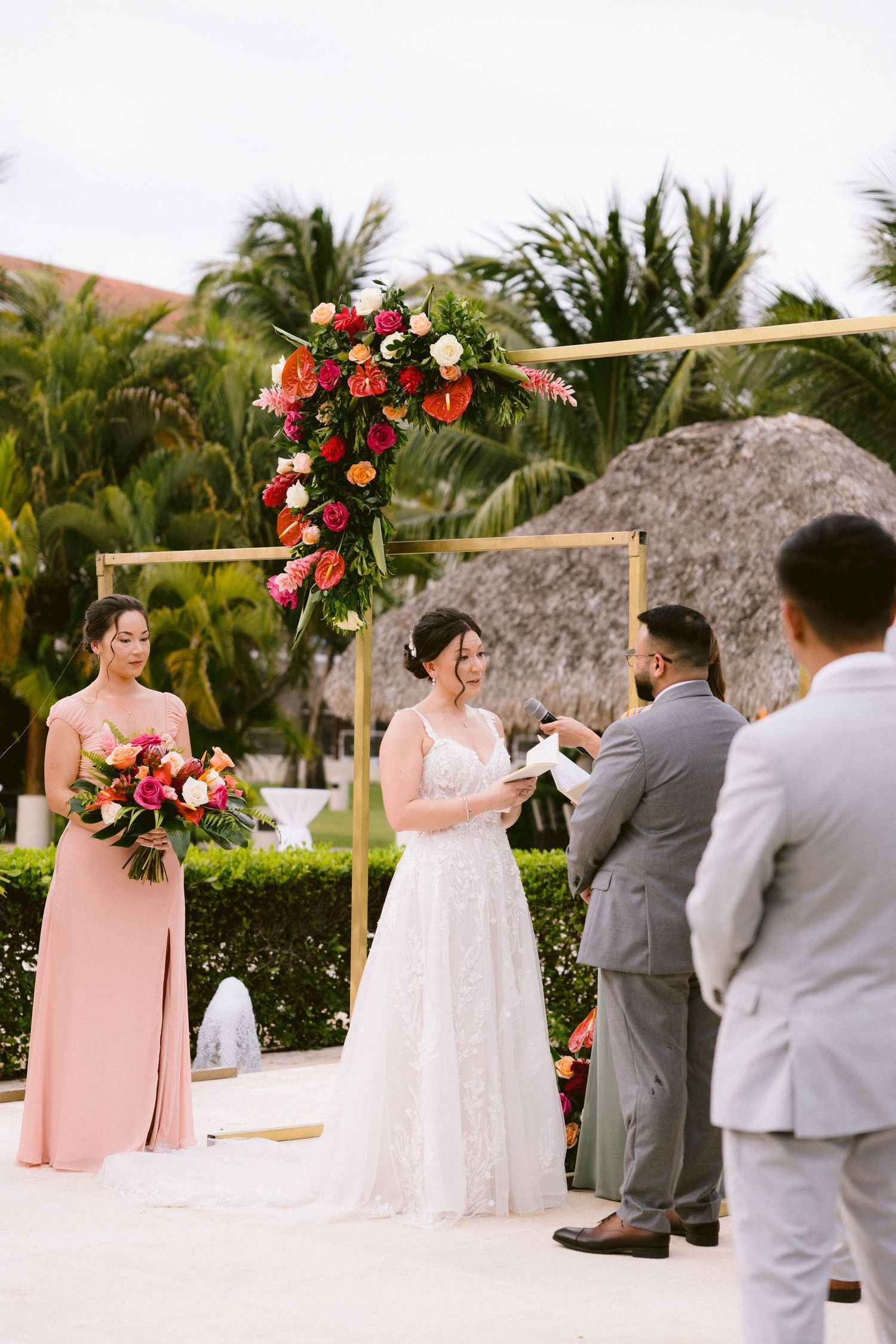 A tropical outdoor wedding ceremony with bamboo arch decorated in red and white flowers, with palm trees in background.