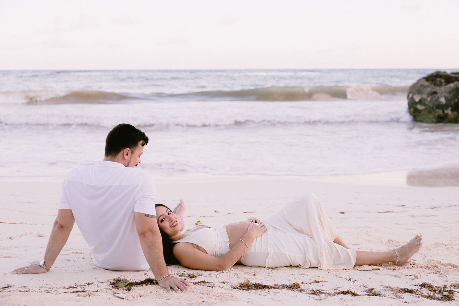 Couple relaxing on a sandy beach, woman lying down in white dress, man sitting beside her near ocean waves.