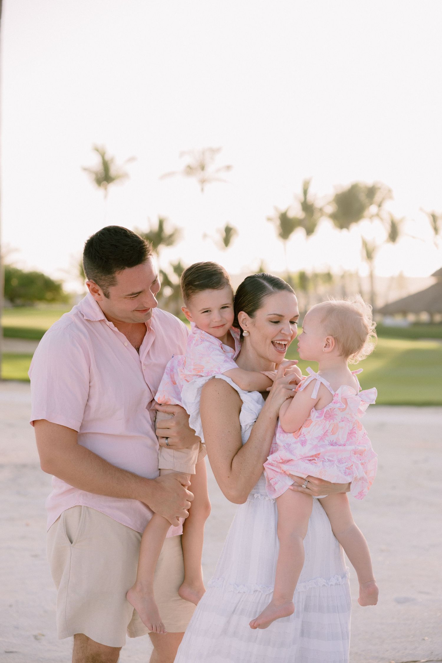 Happy family of four enjoying a sunny beach day, parents holding two young girls in matching pink outfits with palm trees.