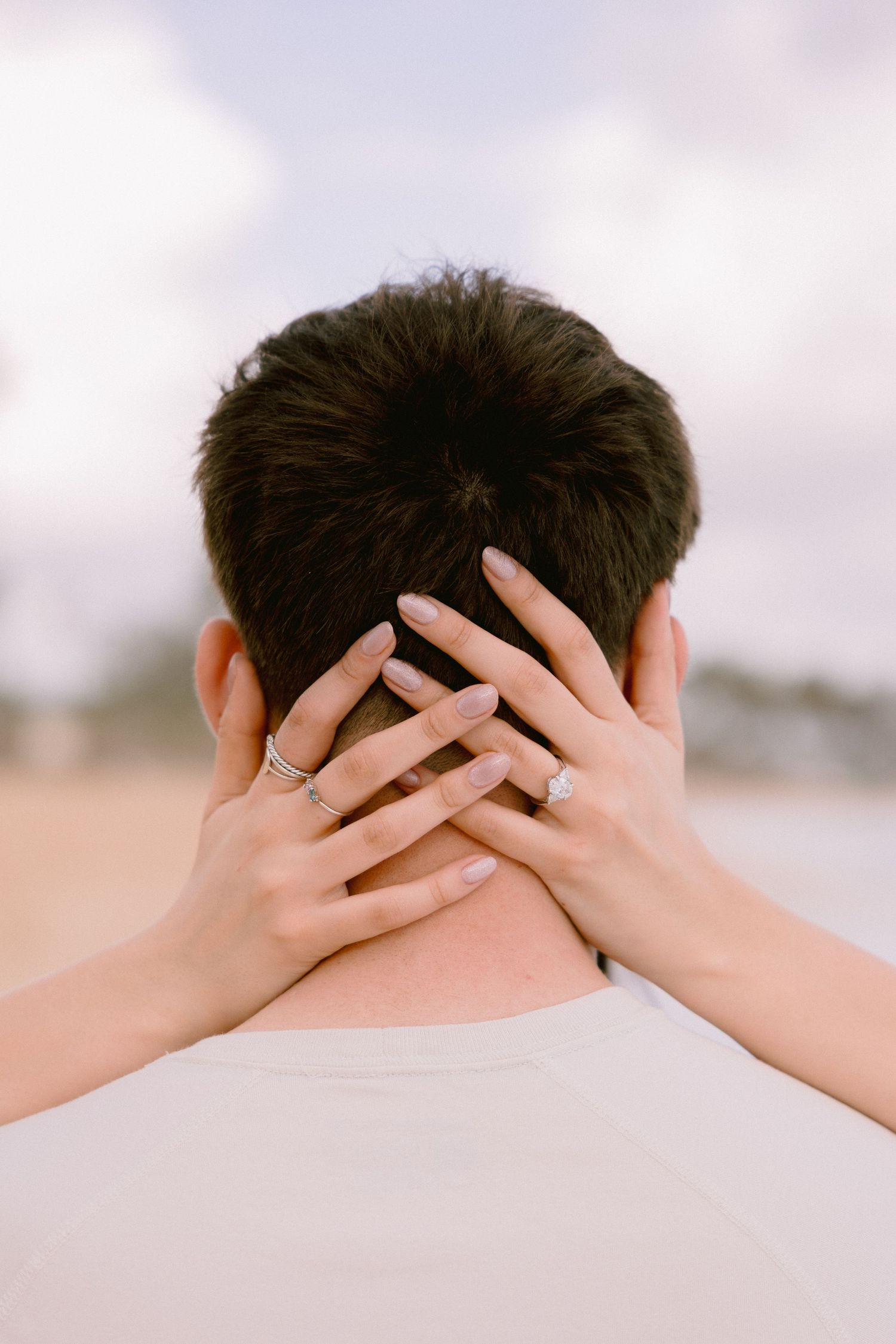 Woman with engagement ring resting hands on back of man's neck at beach, romantic couple moment.