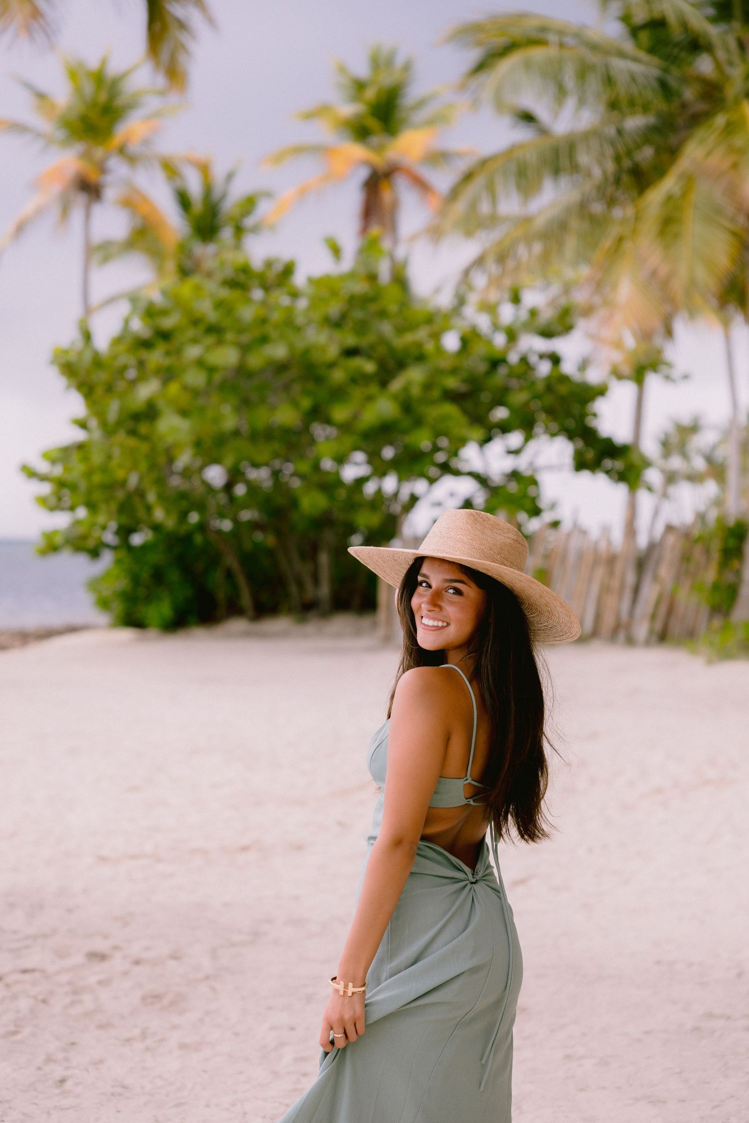 Smiling woman in straw hat and sage green dress poses on tropical beach with palm trees.