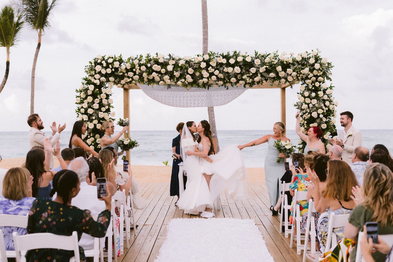 Couple kisses at outdoor beach wedding ceremony under floral arch with white roses, guests celebrating on wooden deck.