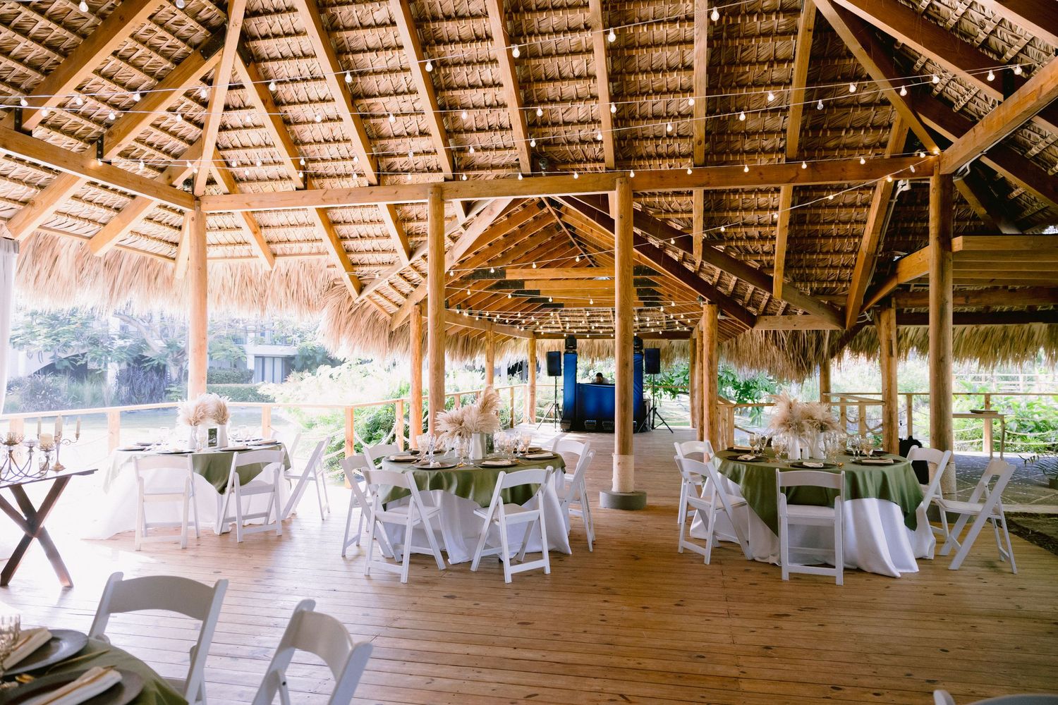 Elegant open-air thatched pavilion wedding reception with white chairs, round tables, and string lights.