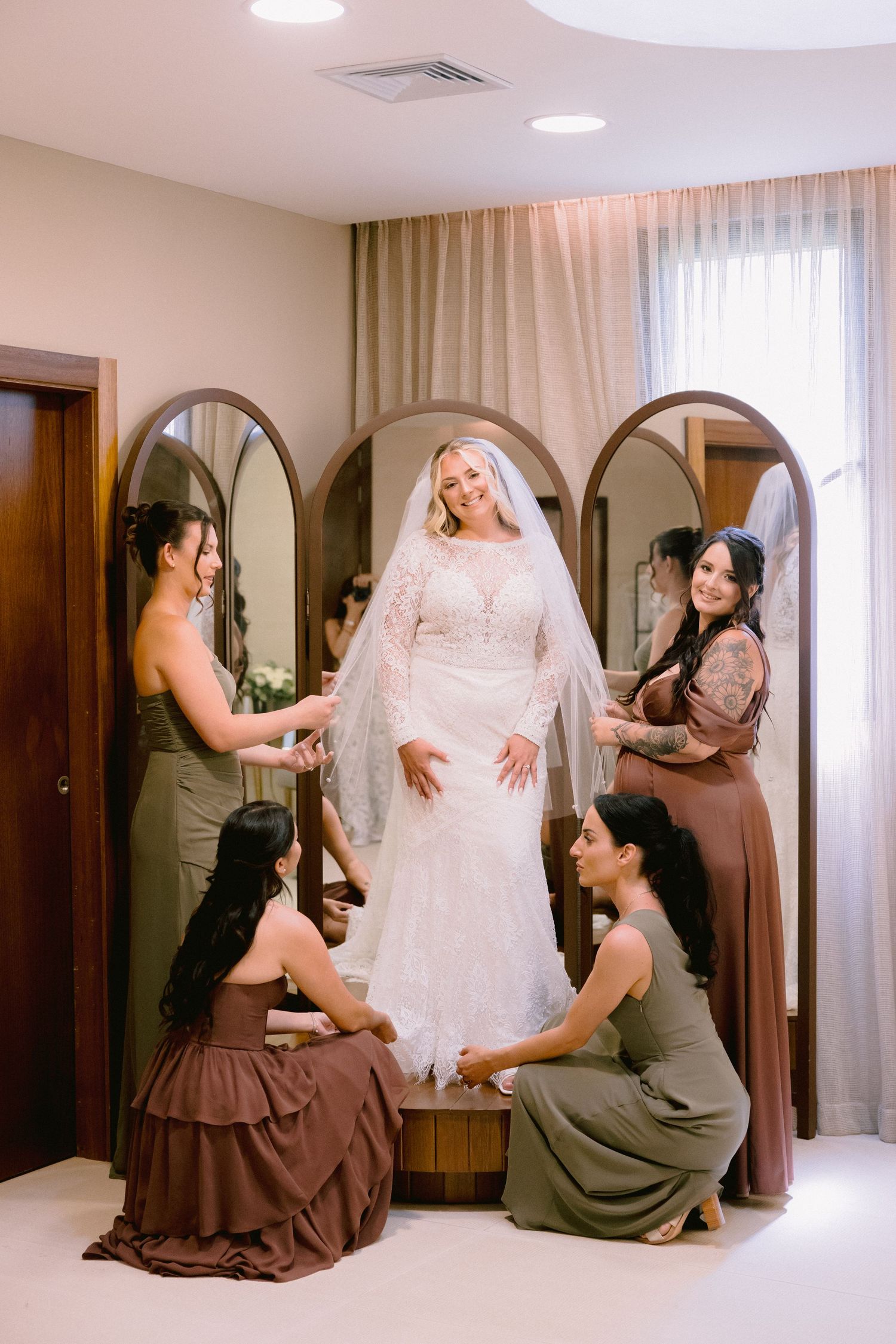 Bride in white lace gown and veil surrounded by bridesmaids in brown dresses helping her get ready.