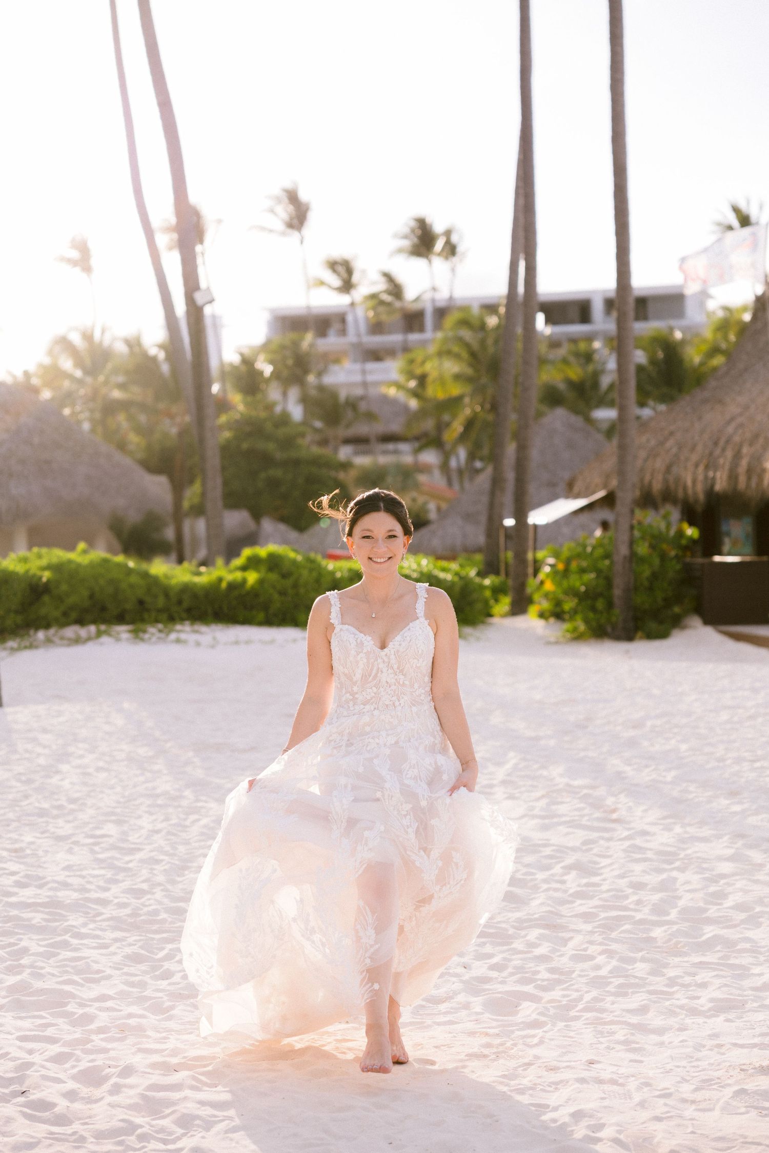 A person in a flowing white wedding dress walks barefoot on a tropical beach with palm trees and thatched huts in background.