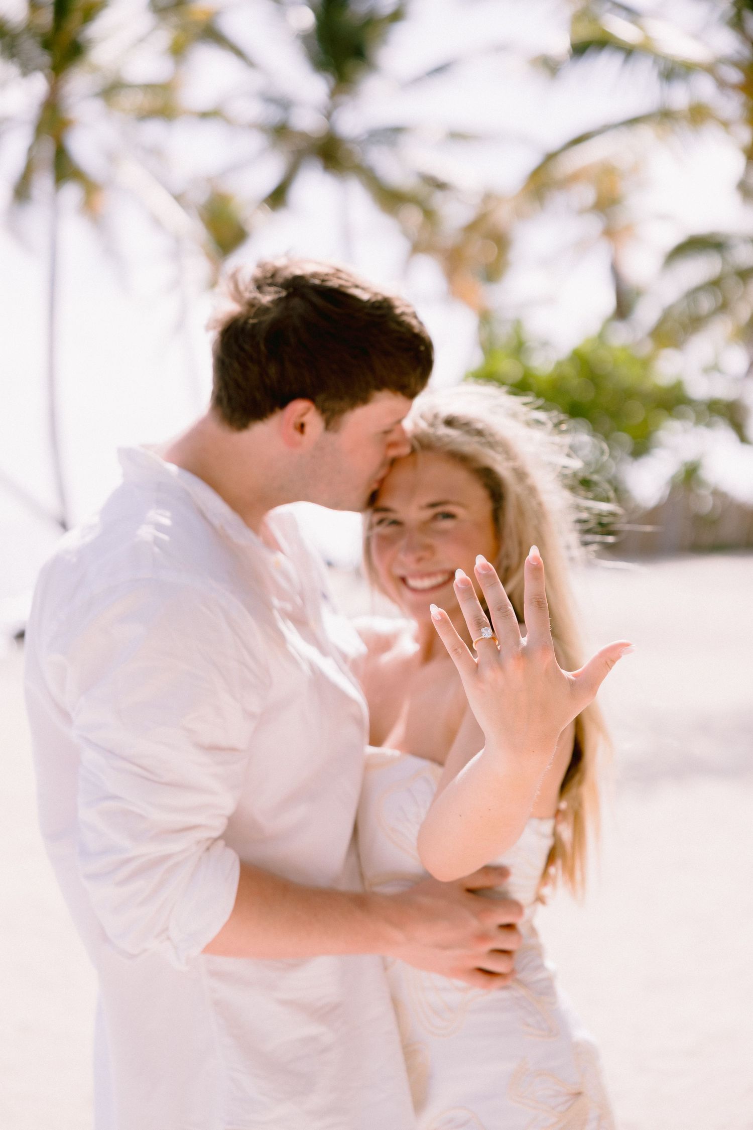 A couple in white outfits embraces lovingly on a tropical beach with palm trees in the background.