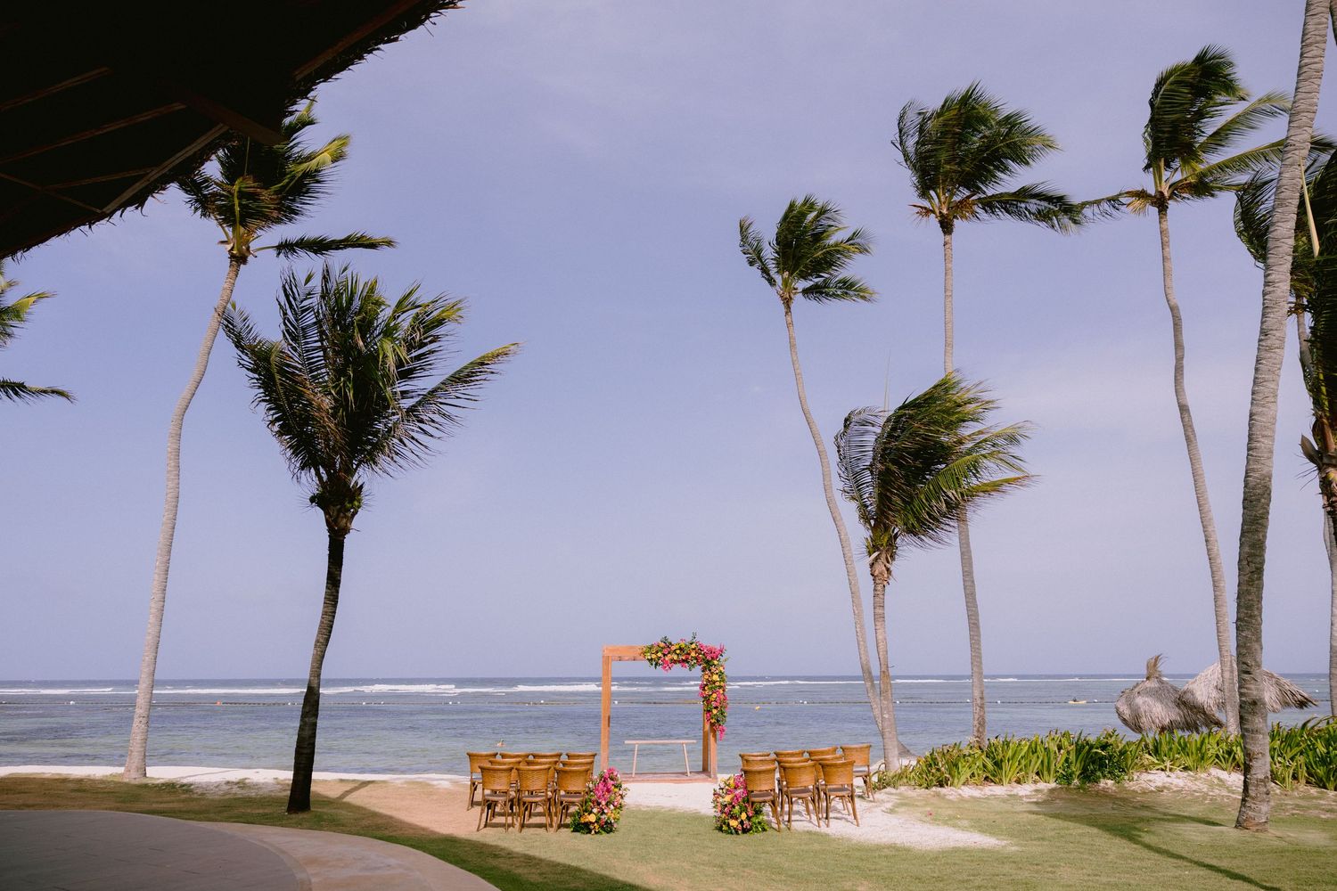 Palm trees sway in strong winds along a tropical beach shoreline with wooden lounge chairs visible in the distance.