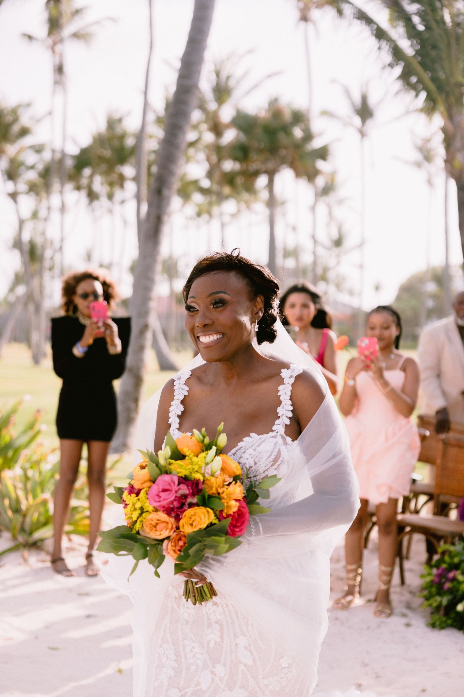A joyful outdoor wedding celebration with tropical palm trees and vibrant orange and pink floral bouquets.
